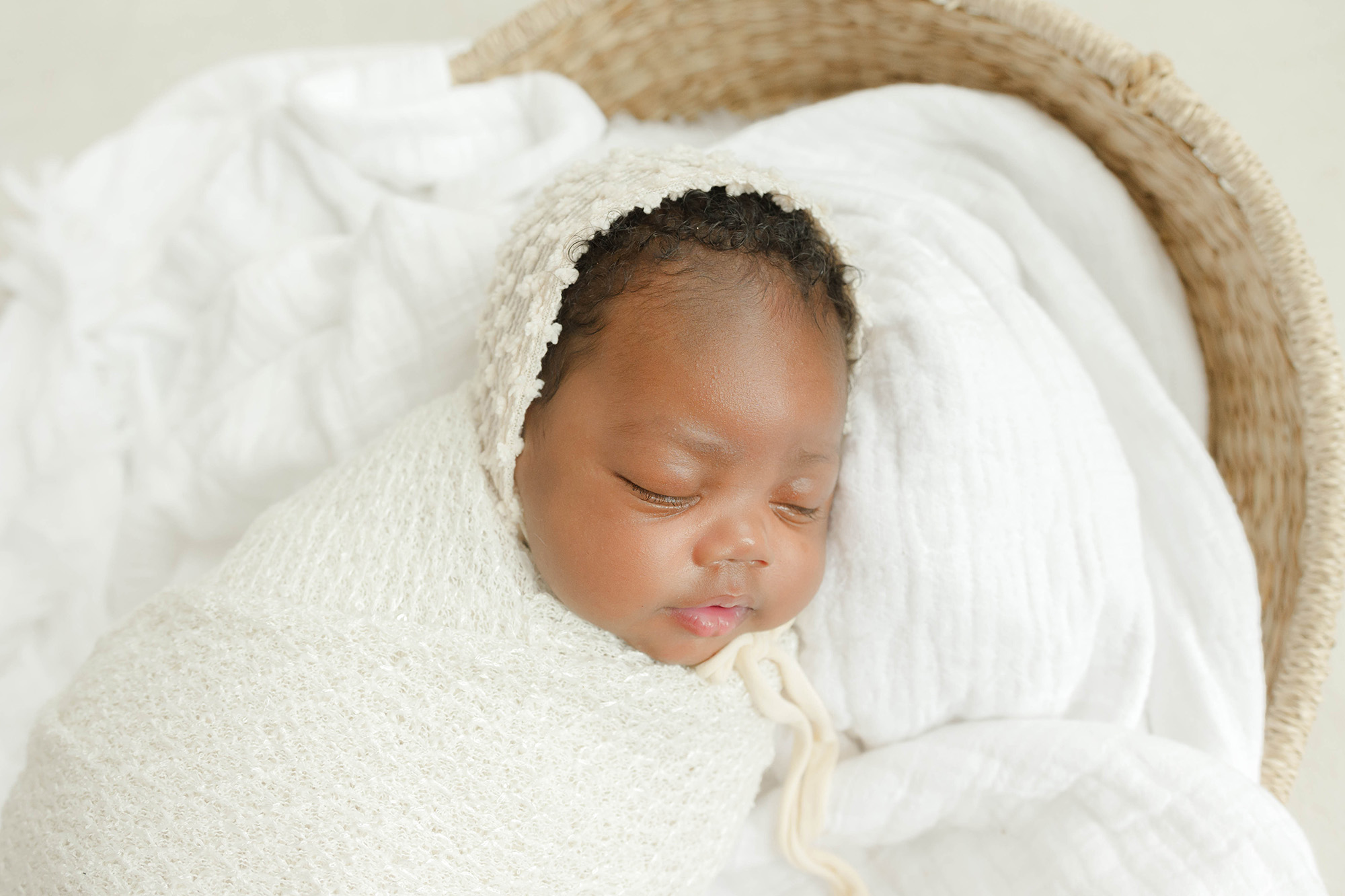 A newborn baby girl sleeps in a white bonnet and swaddle in a woven basket