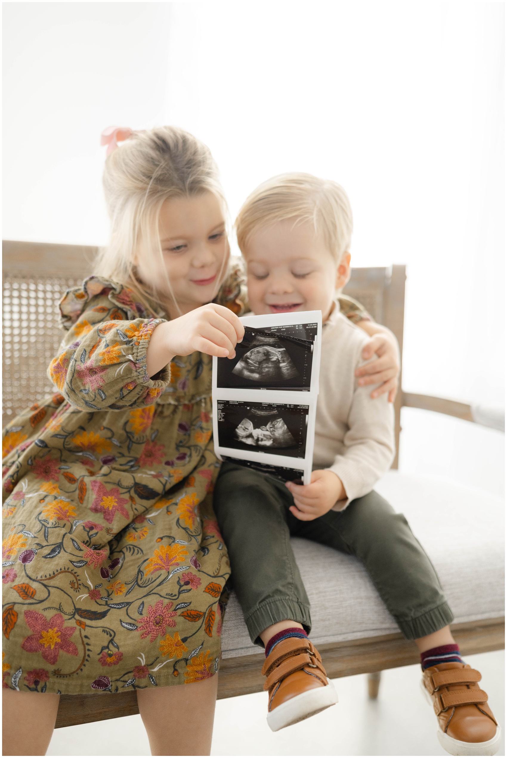 A young girl and her younger brother look at a sonogram while sitting on a bench in a studio