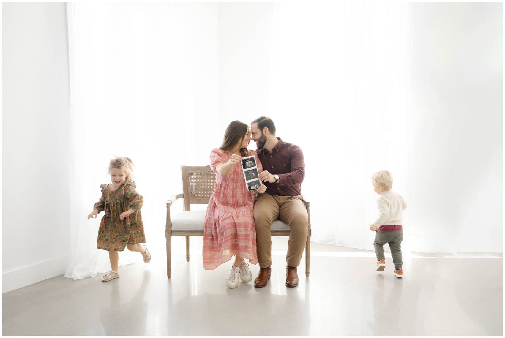 Happy expecting parents sit on a bench showing their sonogram while their two toddlers run around them before visiting a children's museum in okc