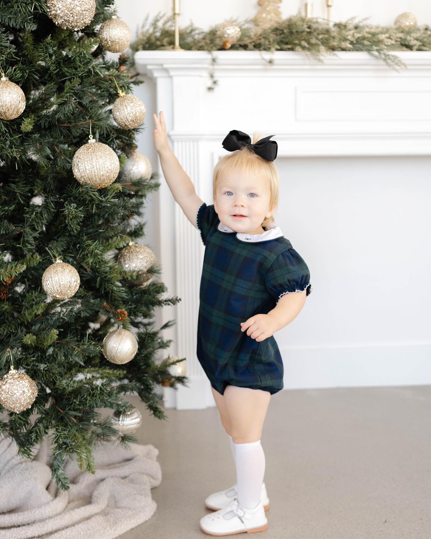 A toddler girl in a plaid dress reaches for an ornament in a studio