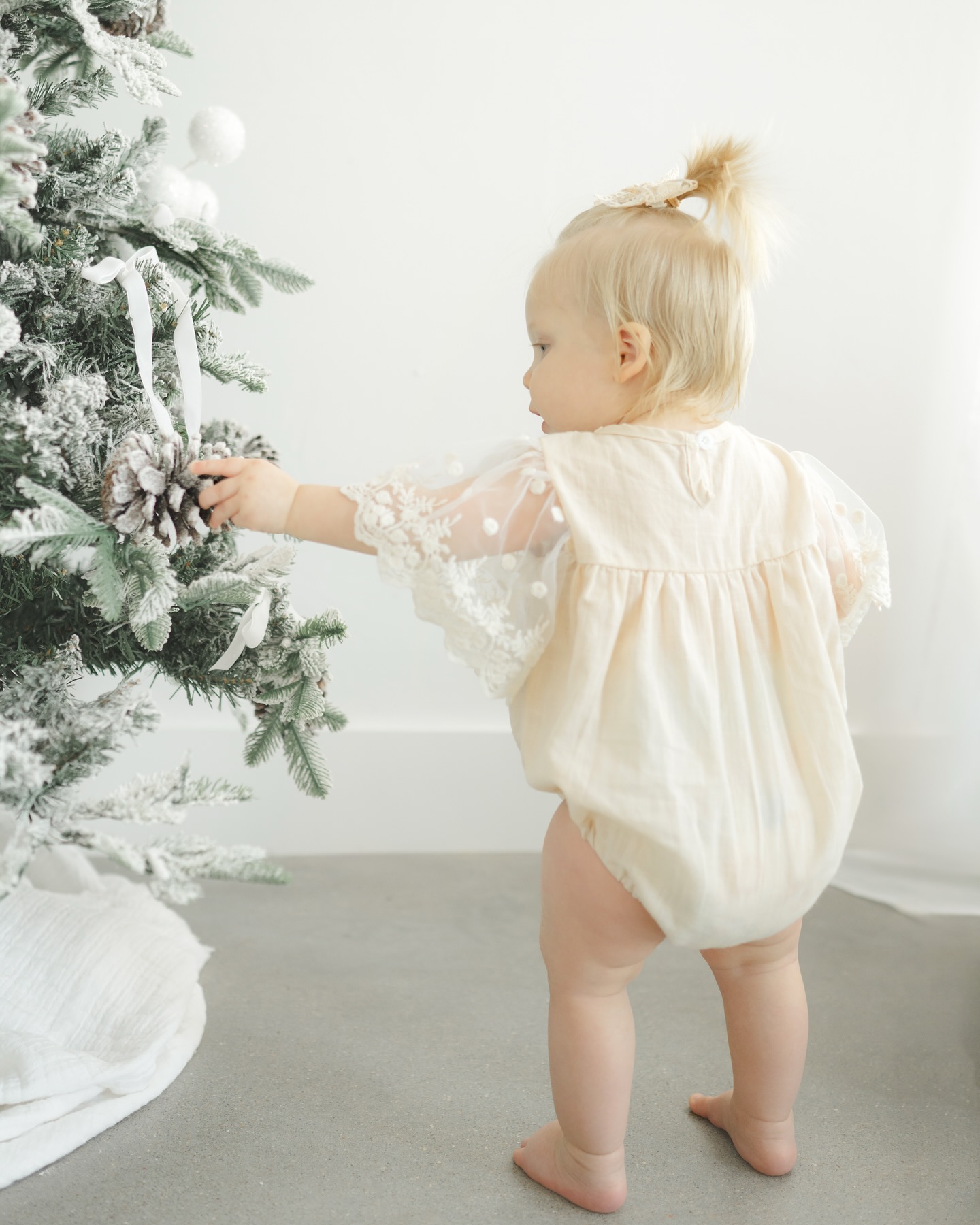 A toddler girl in a yellow onesie explores a pinecone on a tree before visiting a christmas tree farm in okc