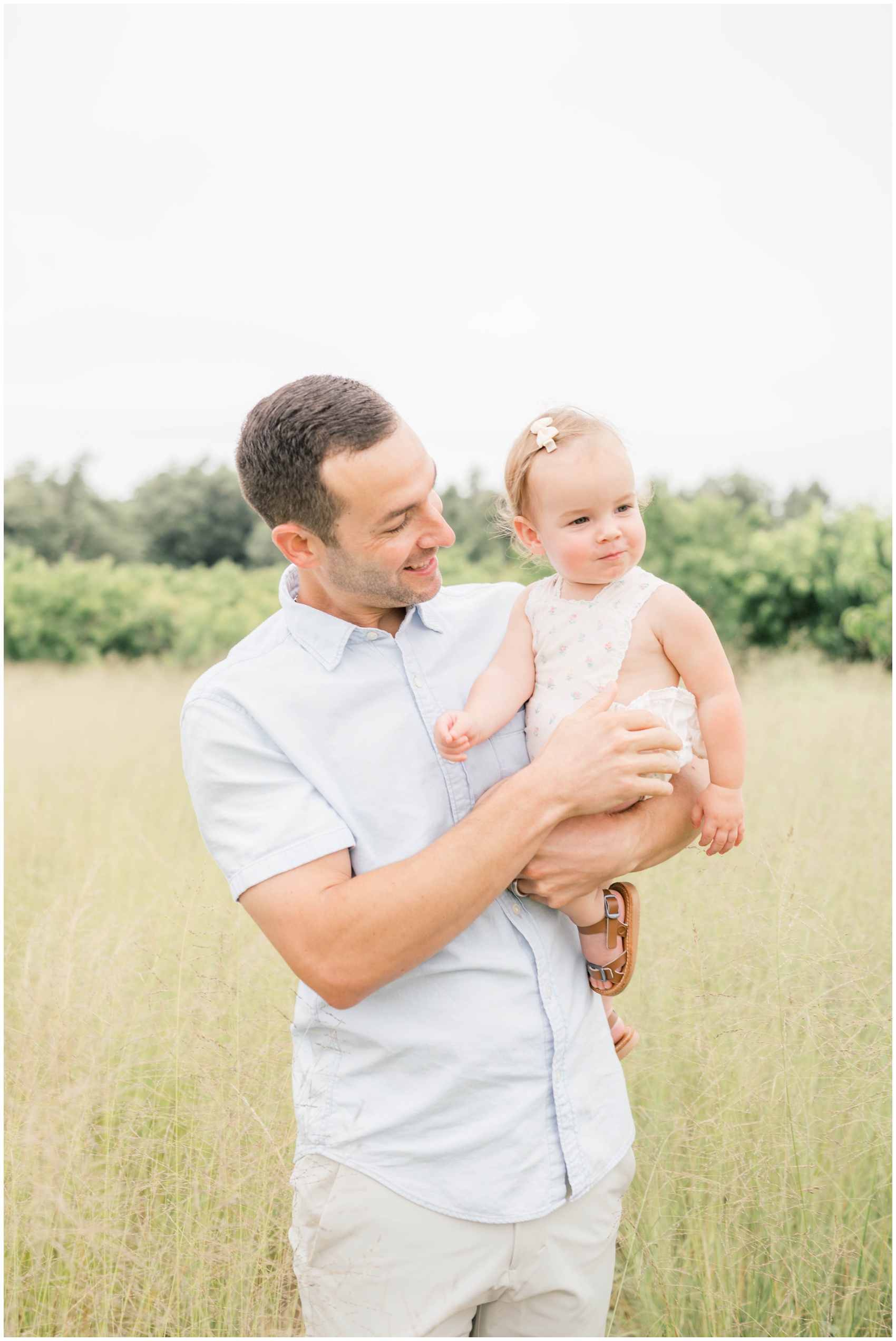 A happy dad smiles at his baby daughter in his arms while standing in a field