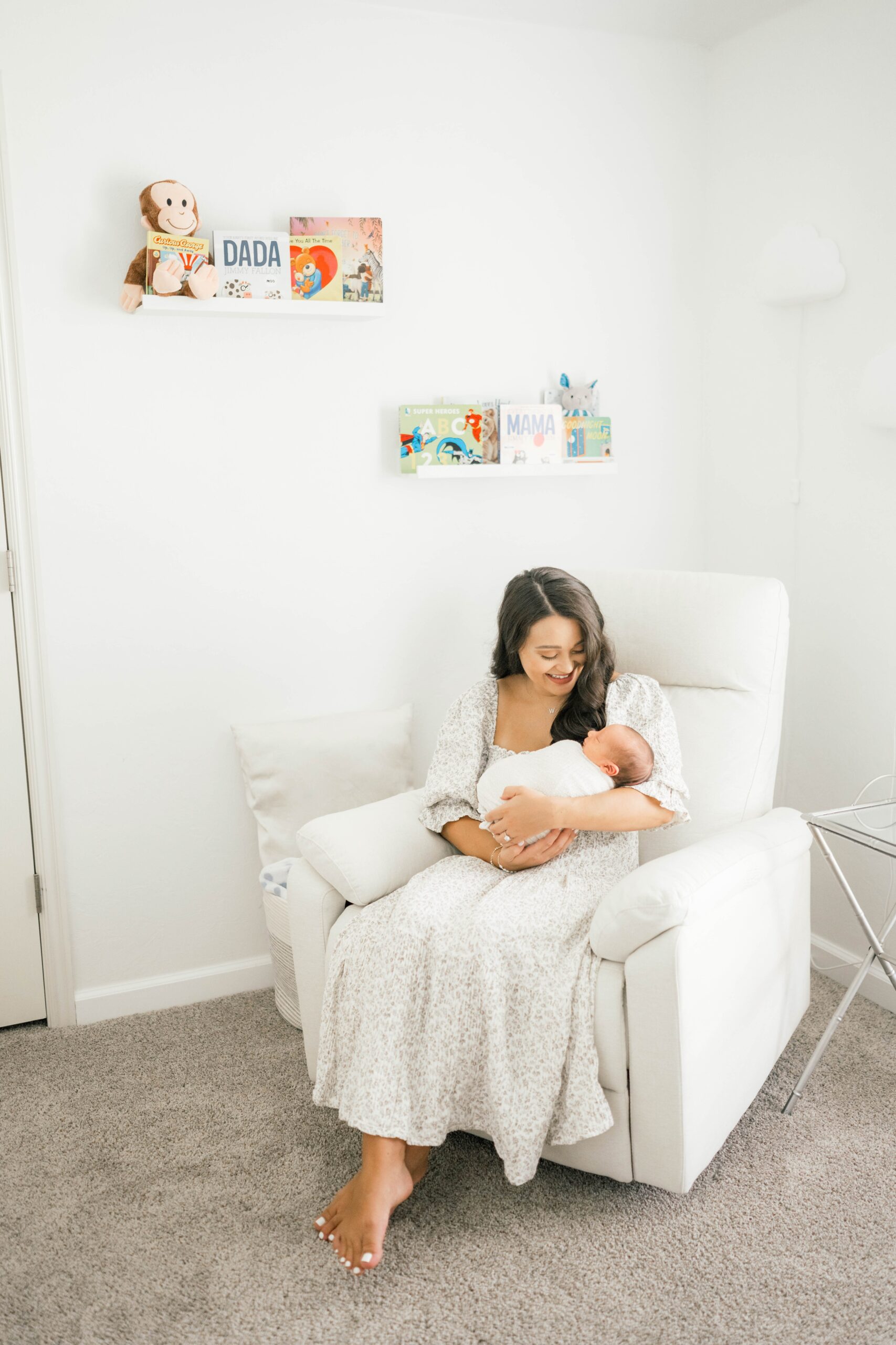 A smiling mom in a white dress sits in a nursery cradling her sleeping newborn