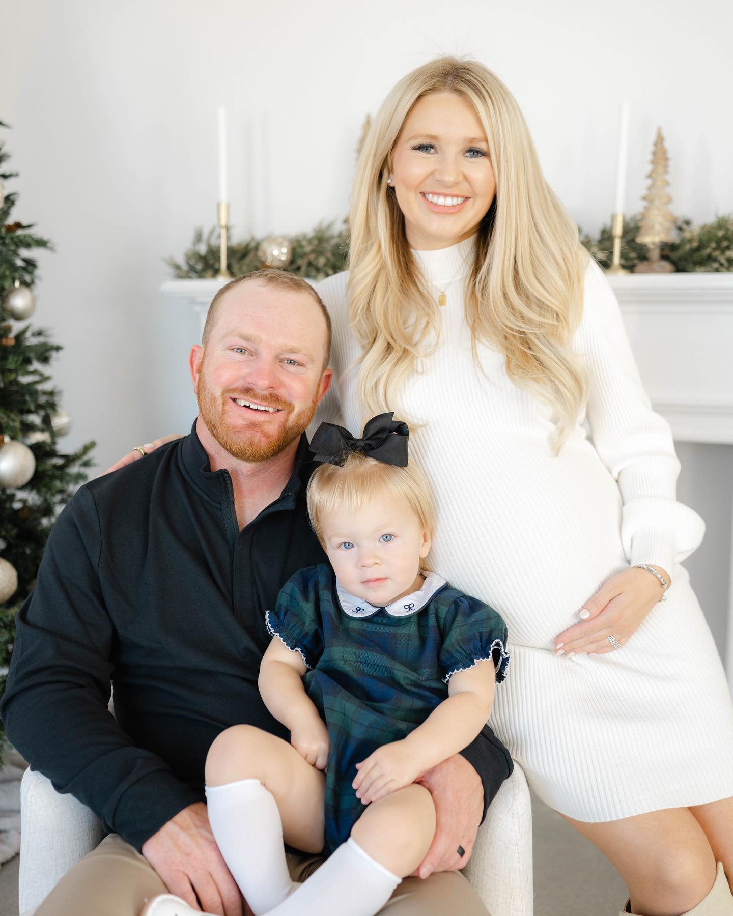 A happy mom and dad sit and lean on a white chair in a christmas living room with their toddler daughter in dad's lap before visiting downtown in december okc