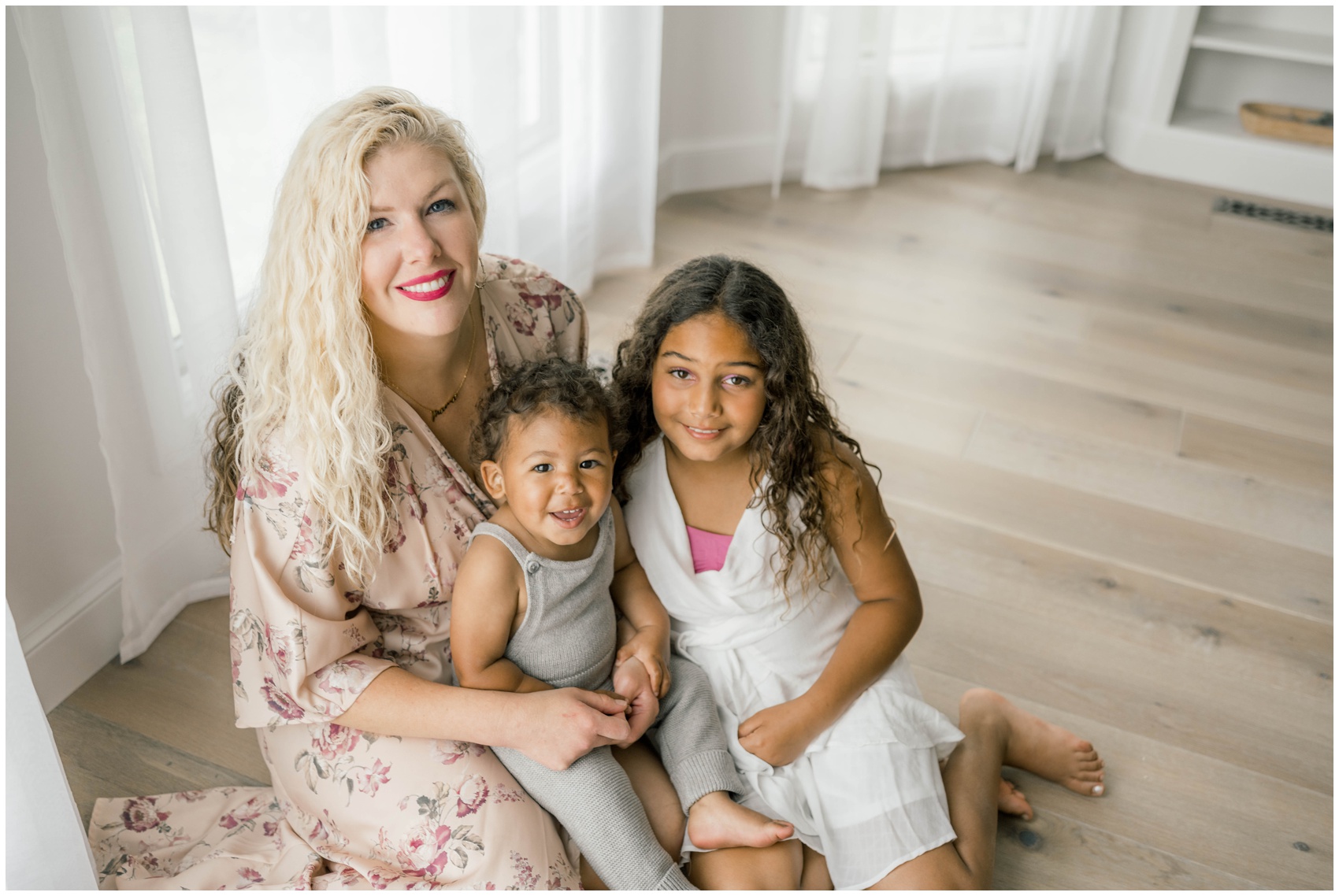 A smiling baby sits in mom's lap on the floor of a studio with big sister