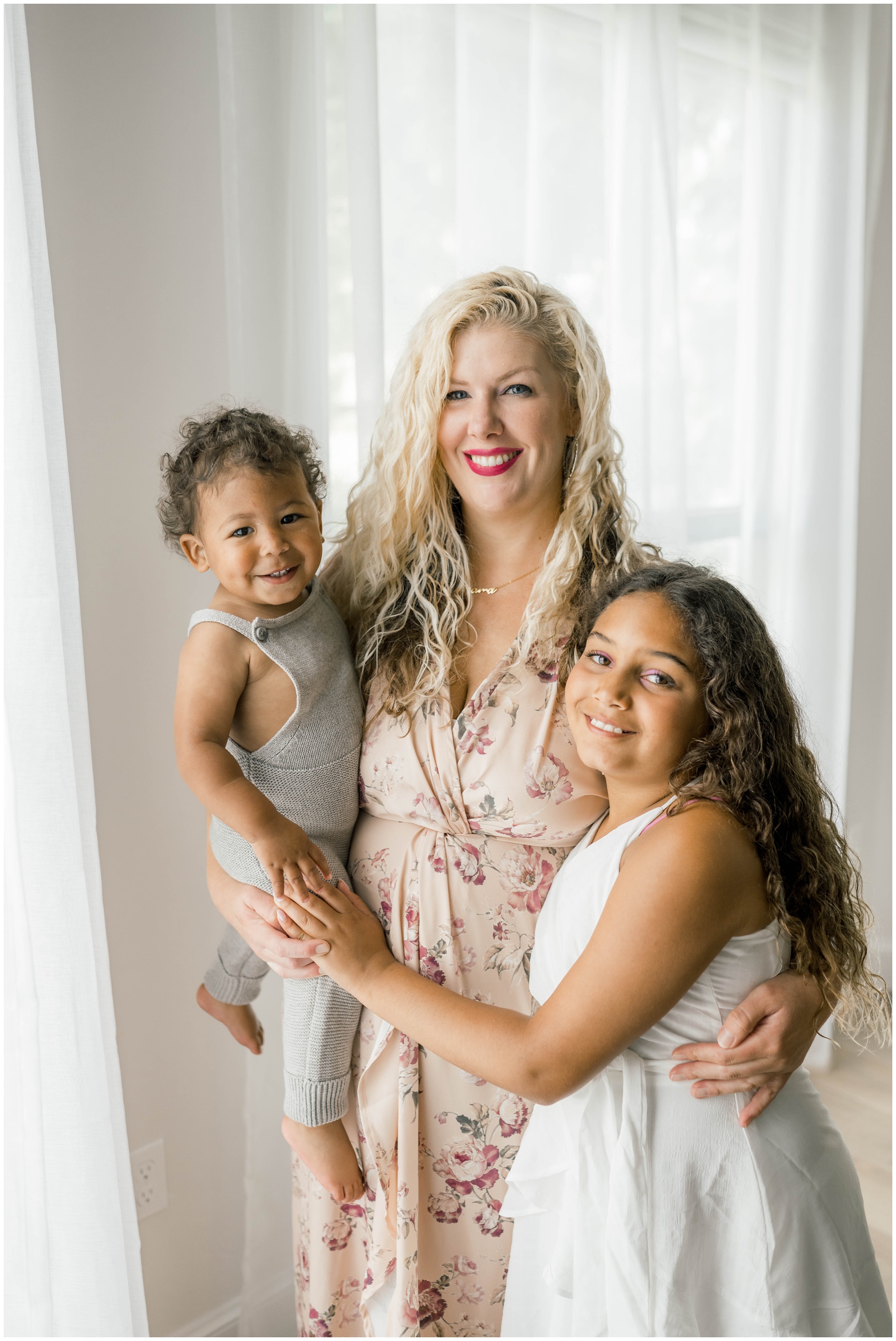 A mother in a floral dress stands hugging her toddler and young daughter in a studio after some family counseling in okc