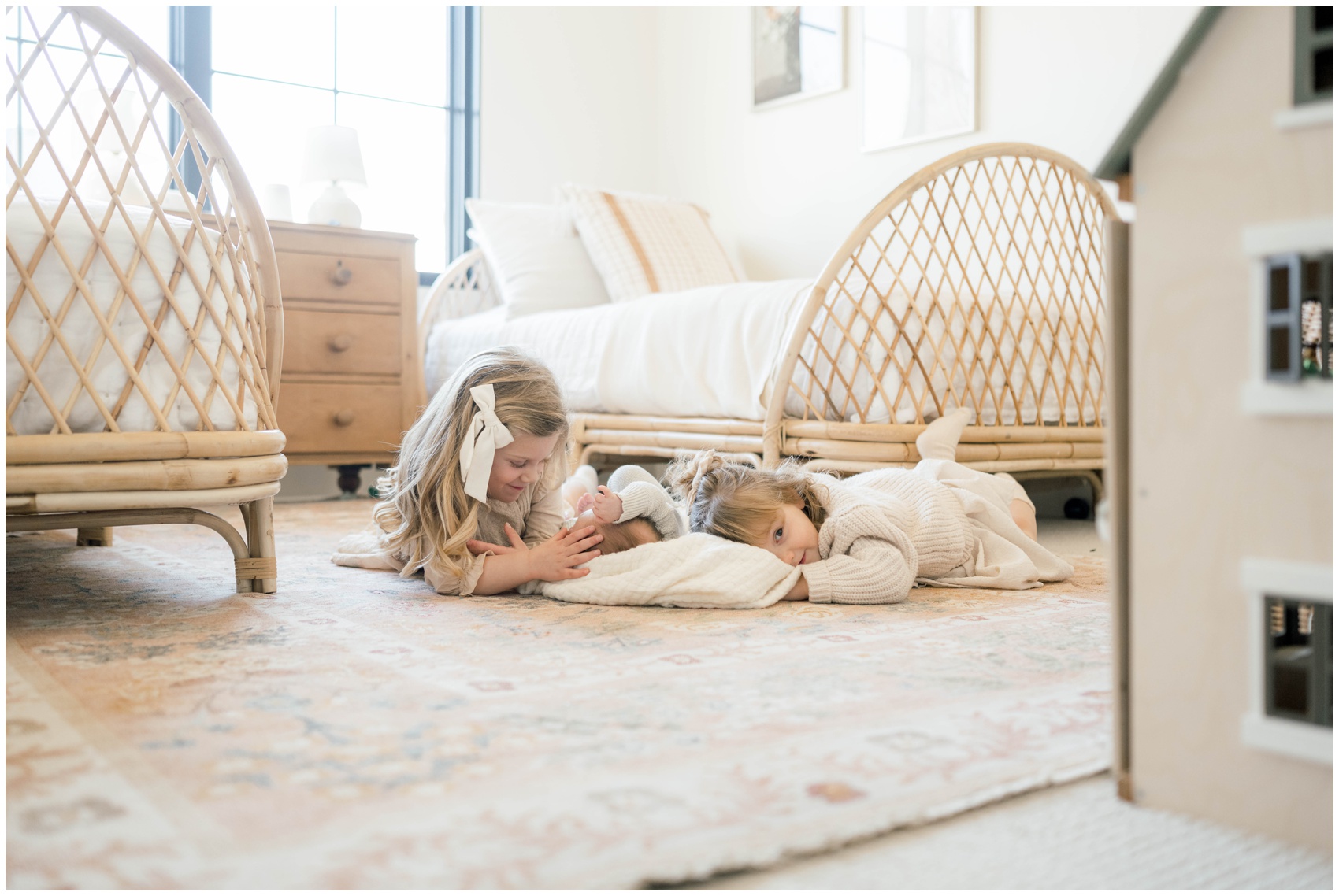 A newborn baby lays on the floor on a rug while its older sisters play with it in their room