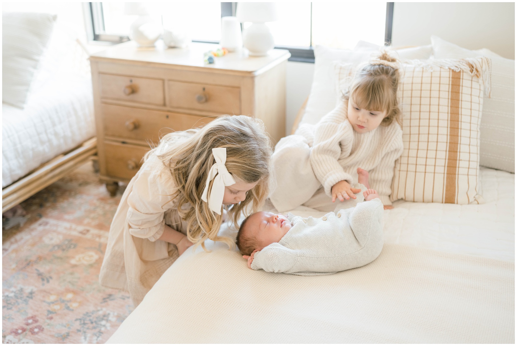 Two young girls in white sweaters explore their sleeping newborn sibling on a bed during family things to do in okc this weekend