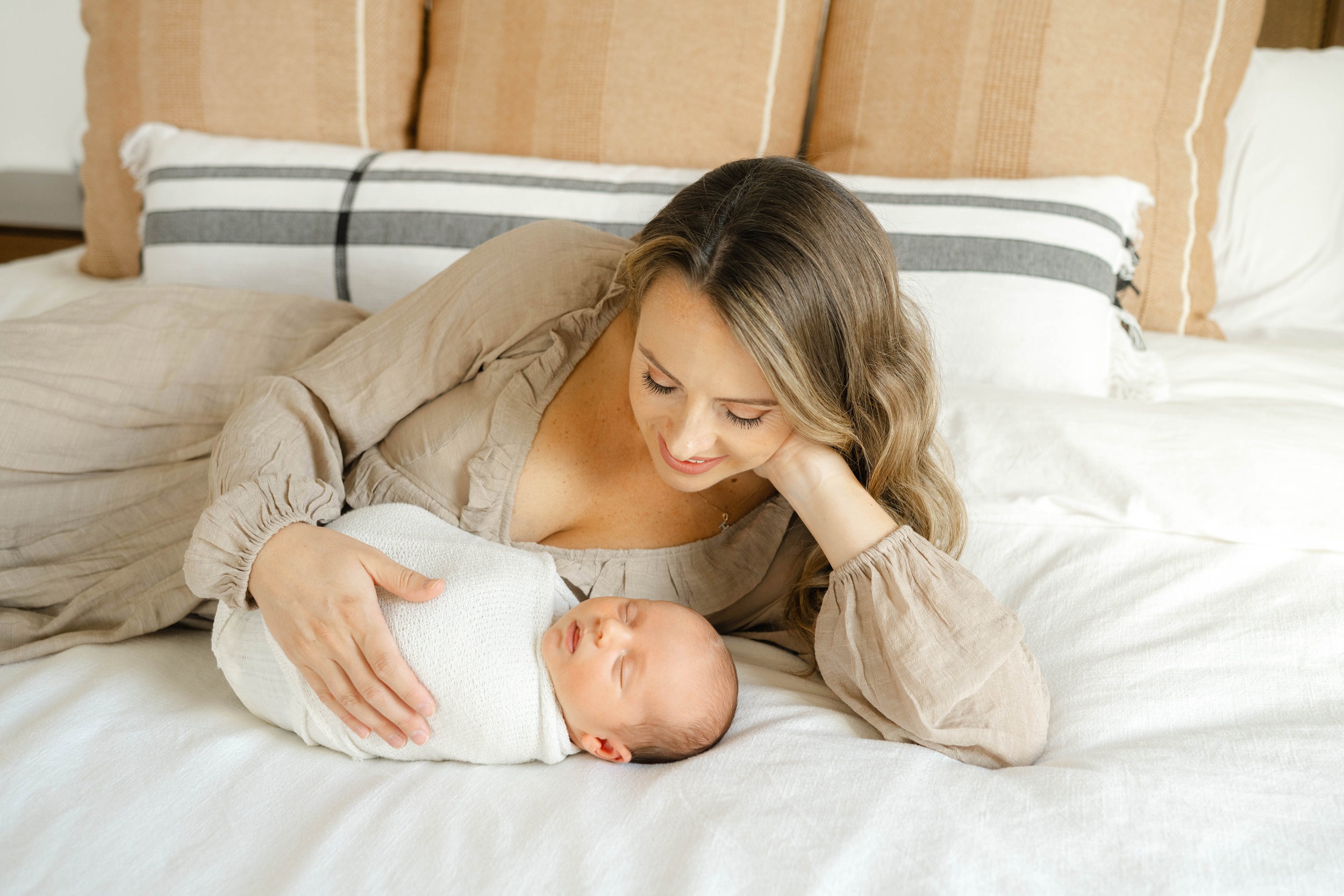 A smiling new mom in a brown dress snuggles her sleeping newborn on a bed