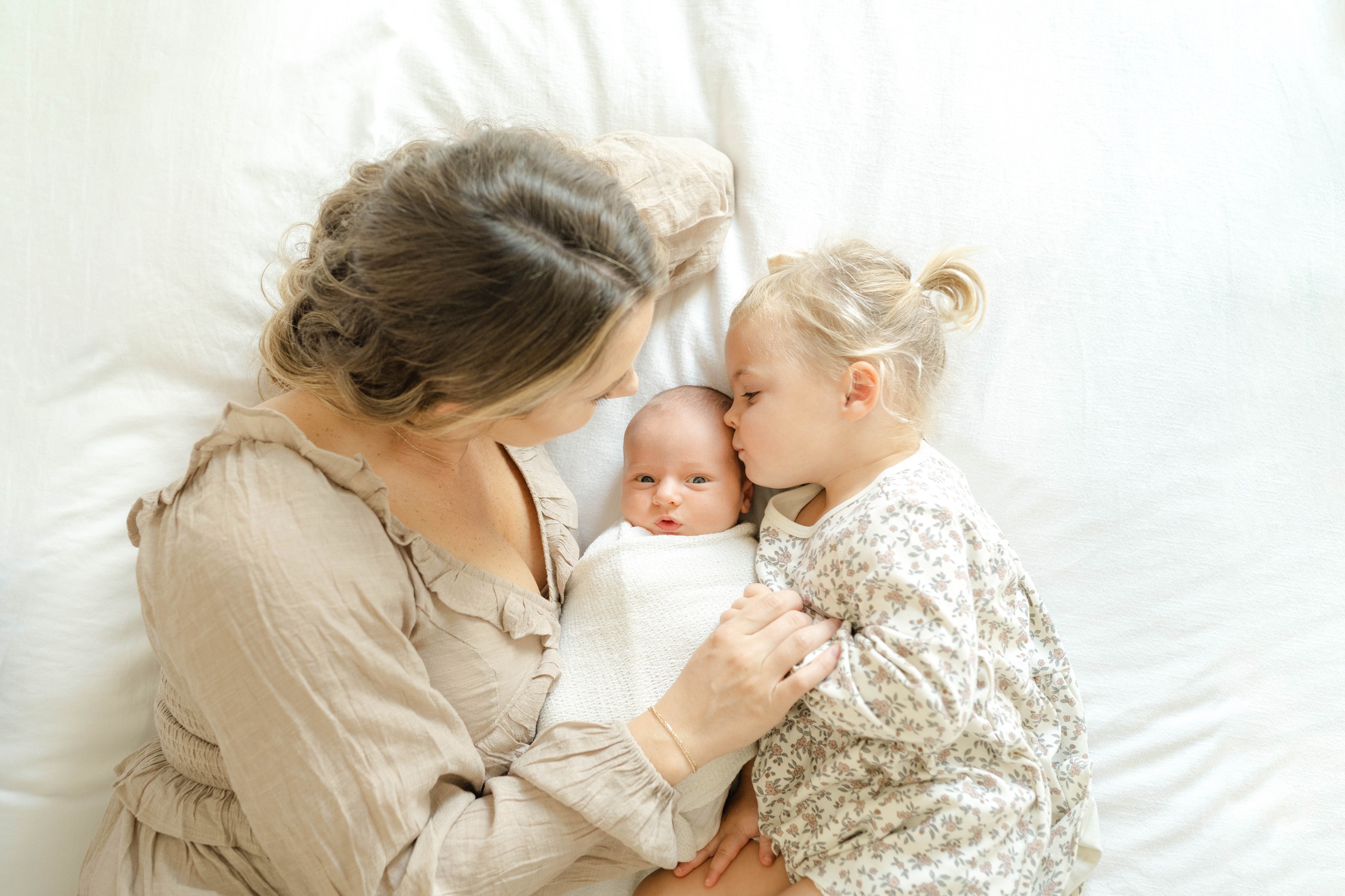 A toddler girl kisses her newborn sibling while cuddling on a bed with mom after visiting fit4mom okc