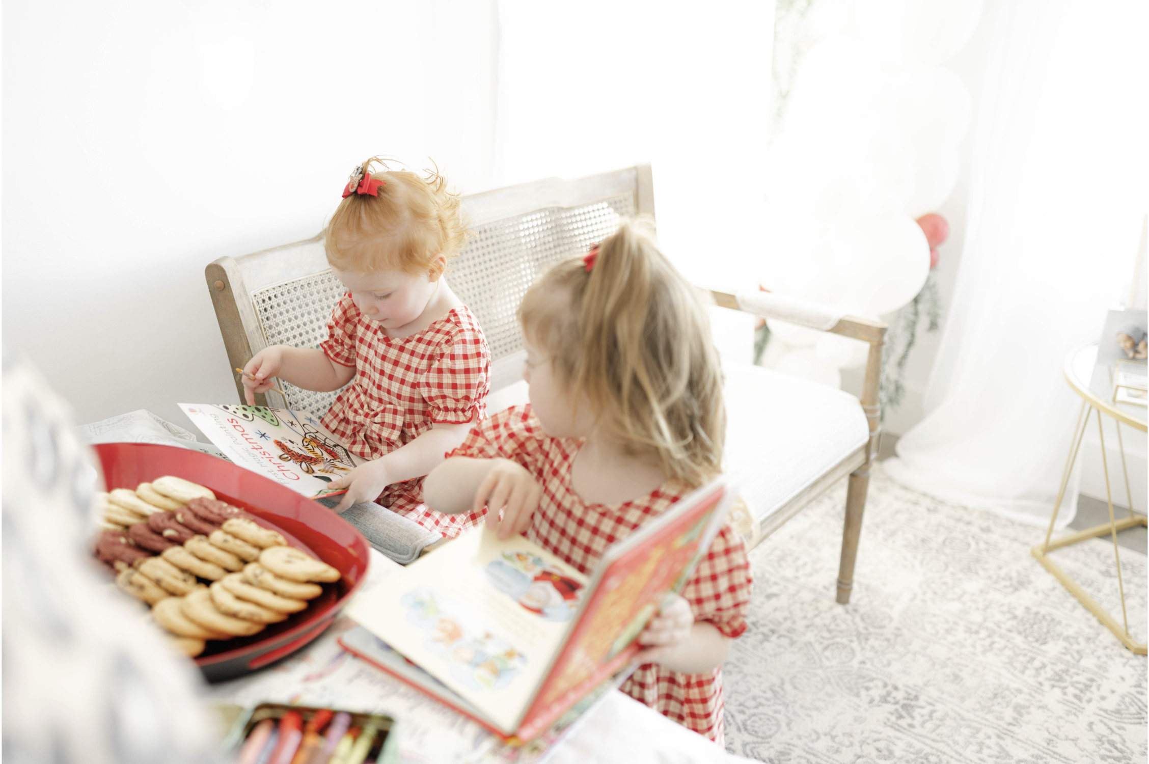 Two toddler girls in matching dresses read book on a bench by a plate of cookies