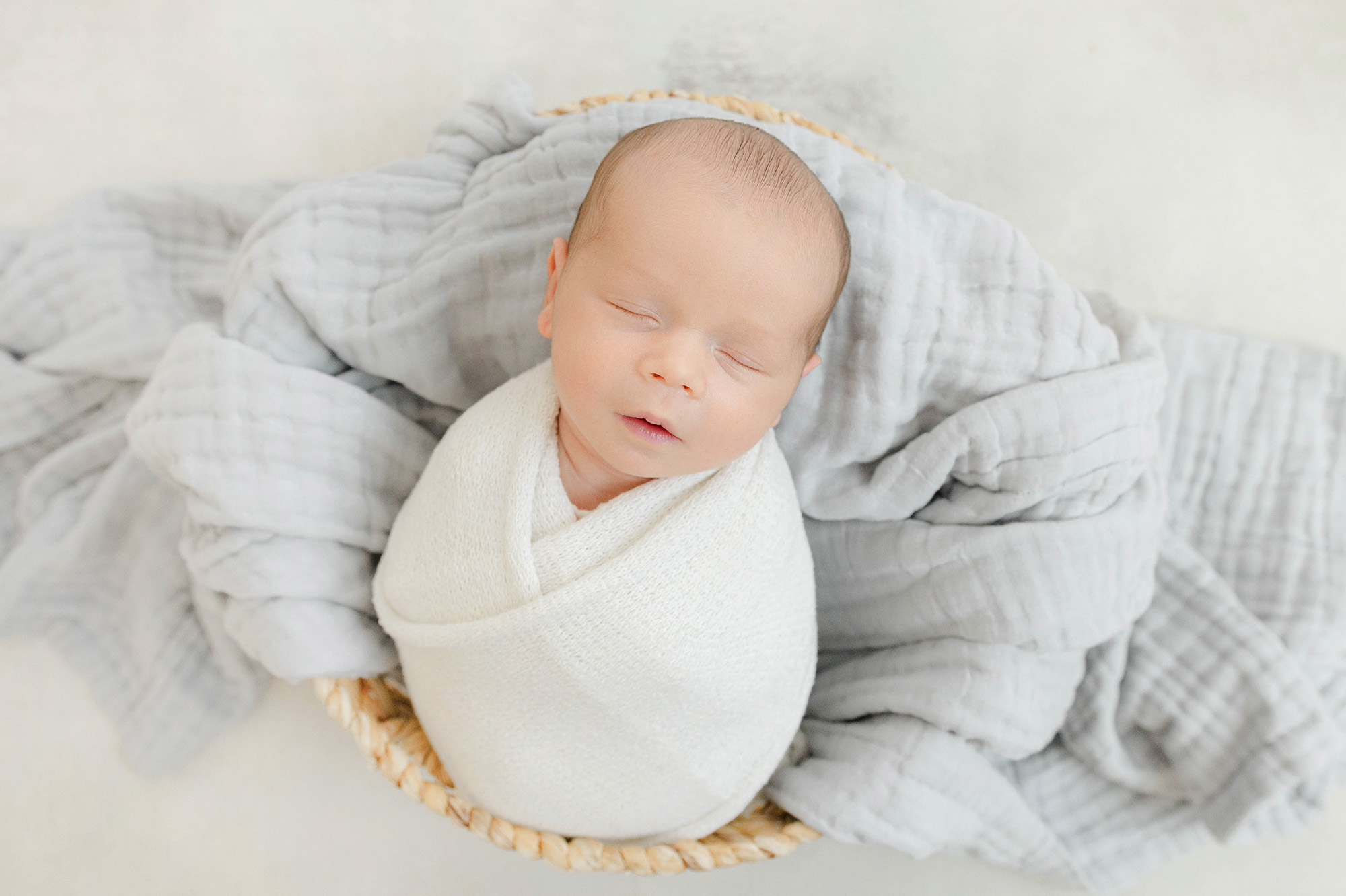 A newborn baby sleeps in a white swaddle in a woven basket