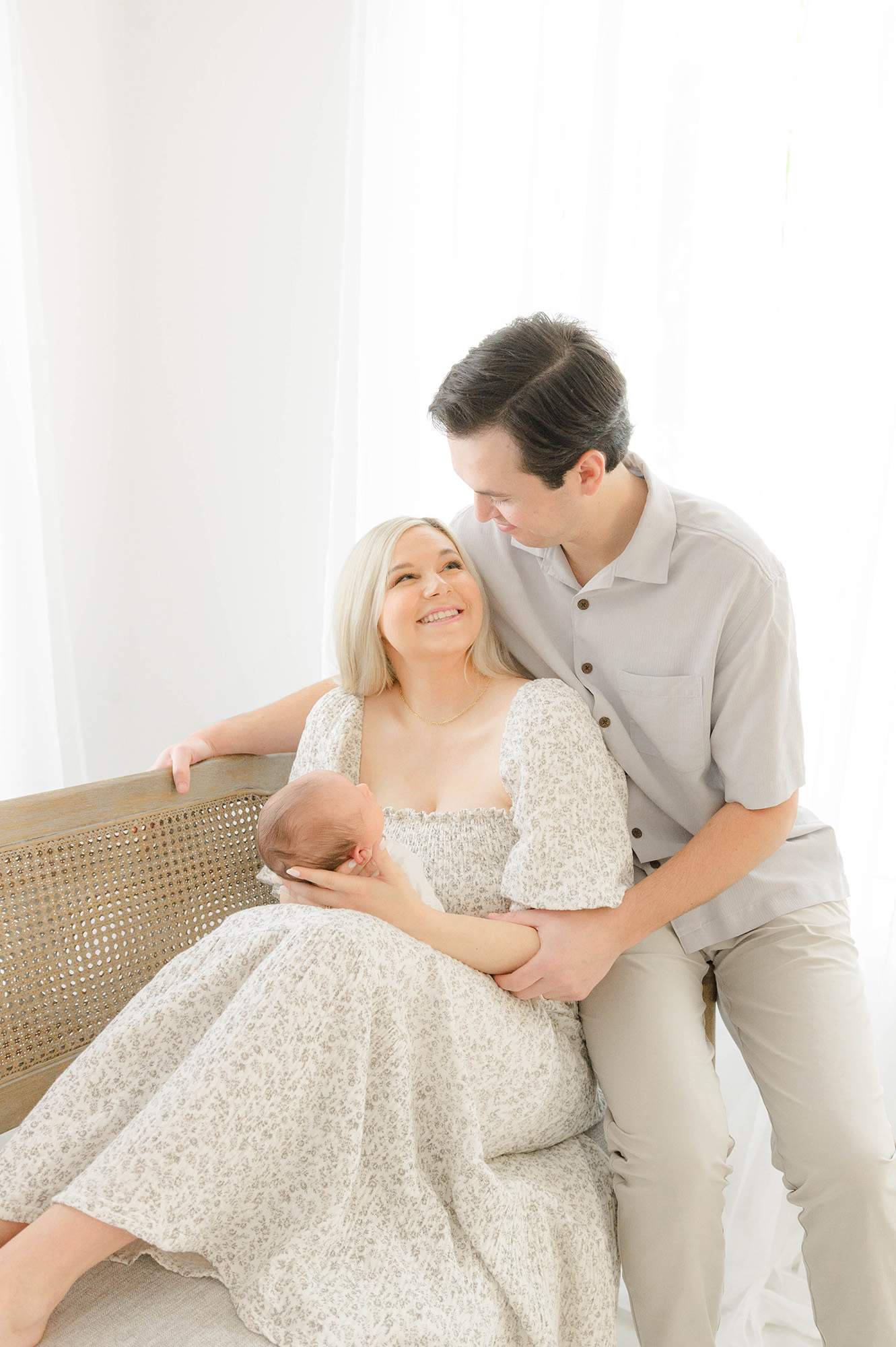 Happy parents sit across a bench in a studio smiling at each other with their newborn in mom's lap after meeting jenni jenkins