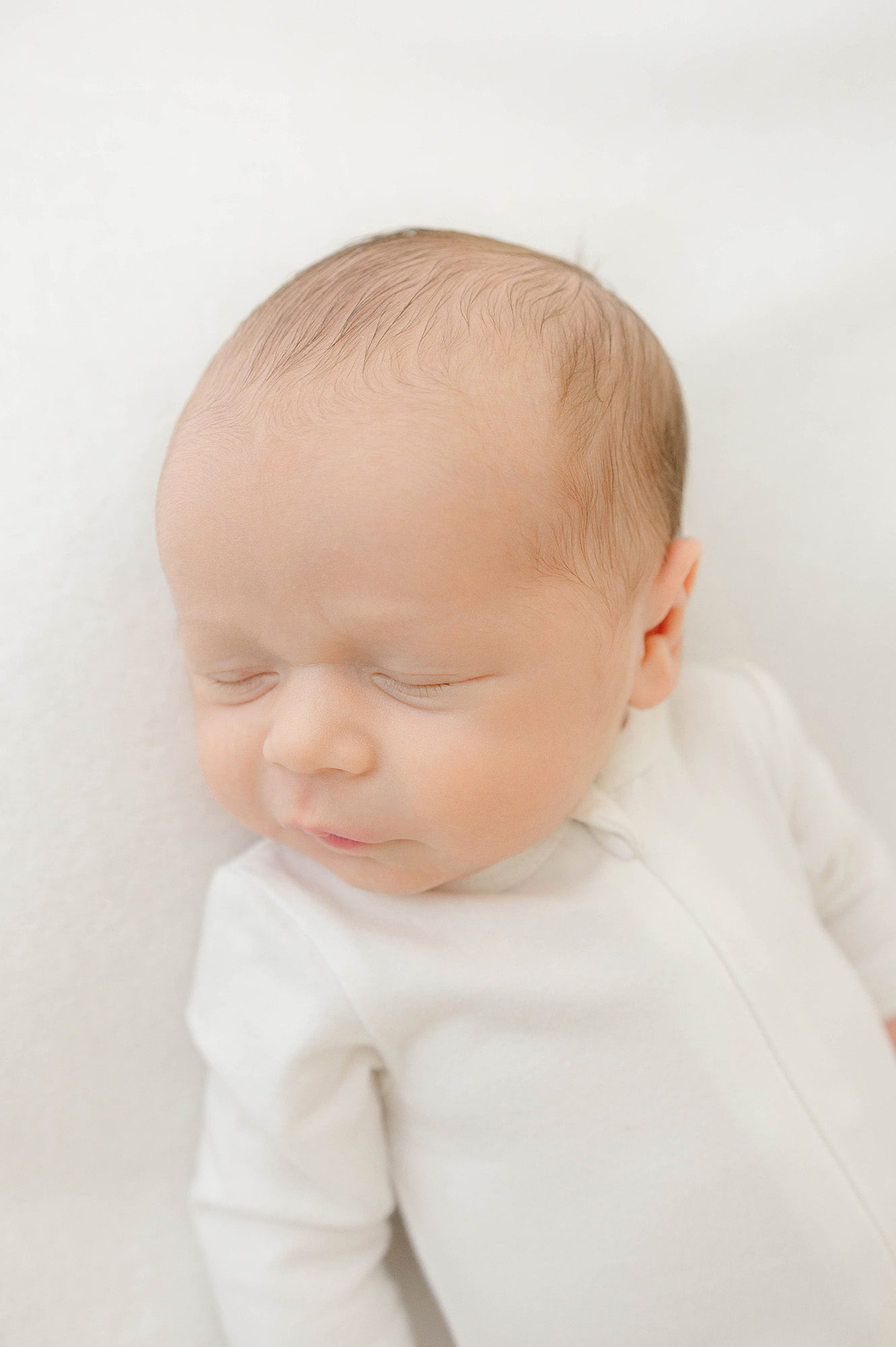 A newborn baby sleeps on a bed in a white onesie on its back thanks to jenni jenkins