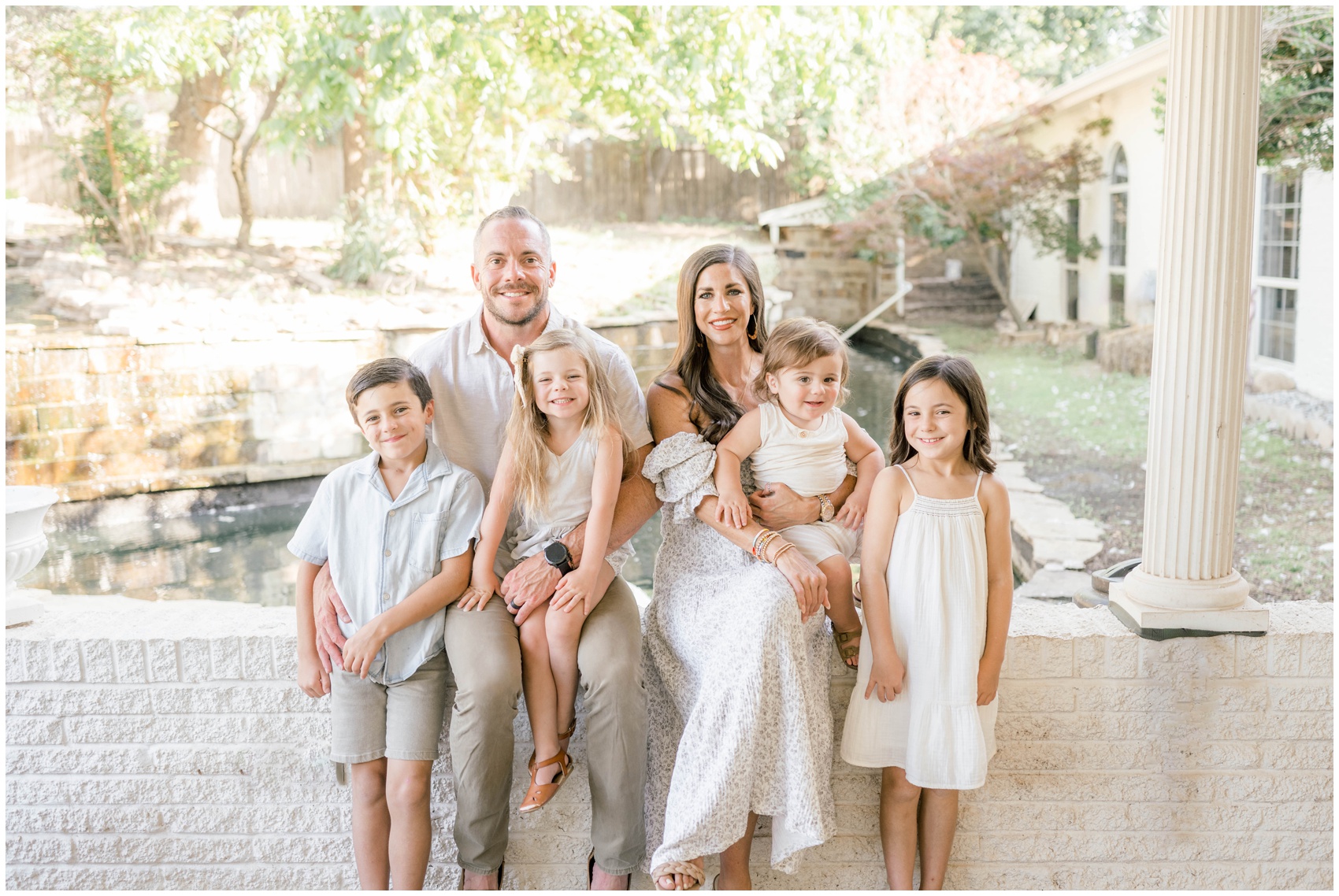 A happy family of 6 with all young children sit on a garden wall smiling and hugging