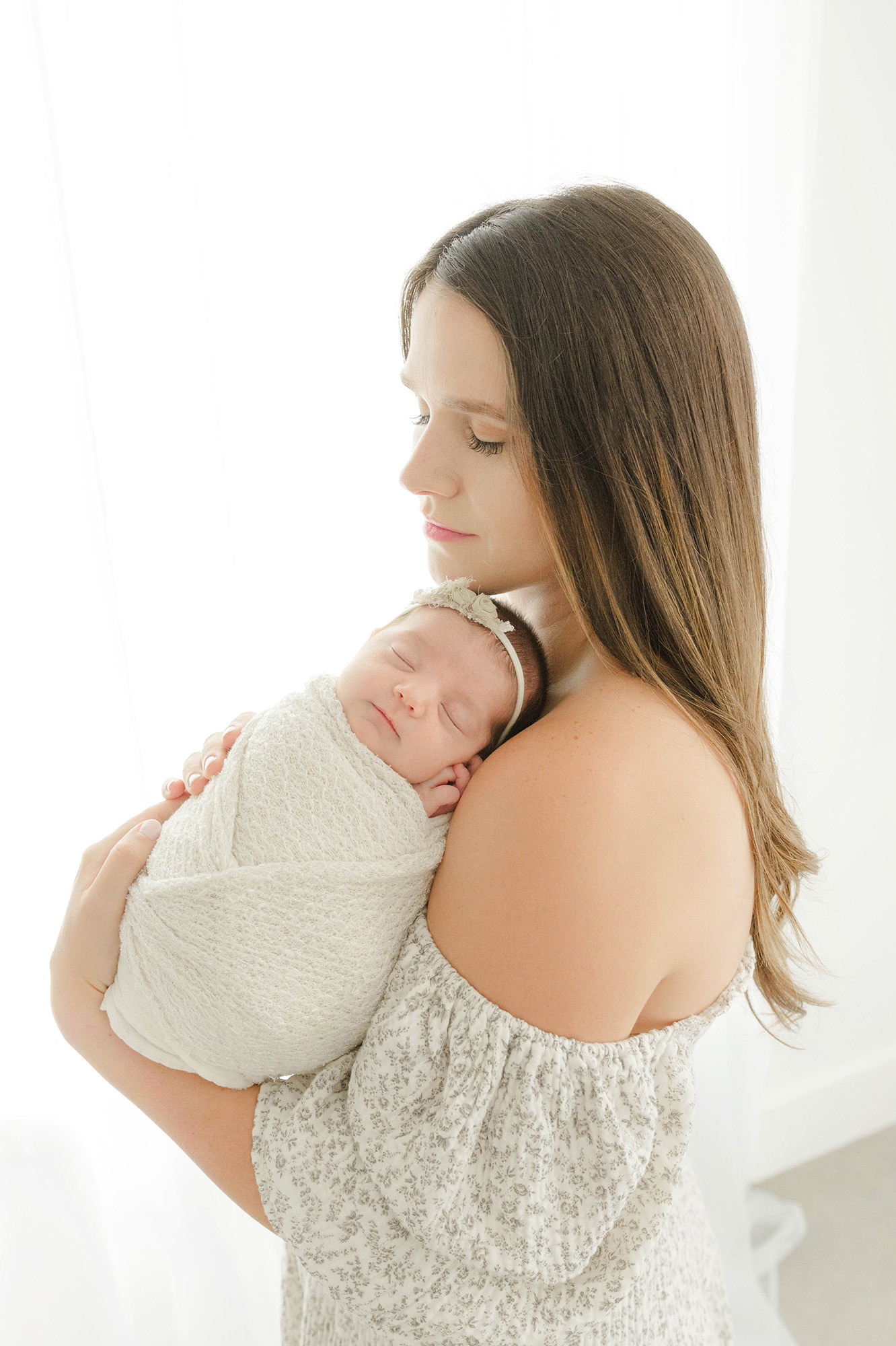 A newborn baby sleeps on mom's shoulder while standing in a studio in a white swaddle thanks to labor of love midwifery