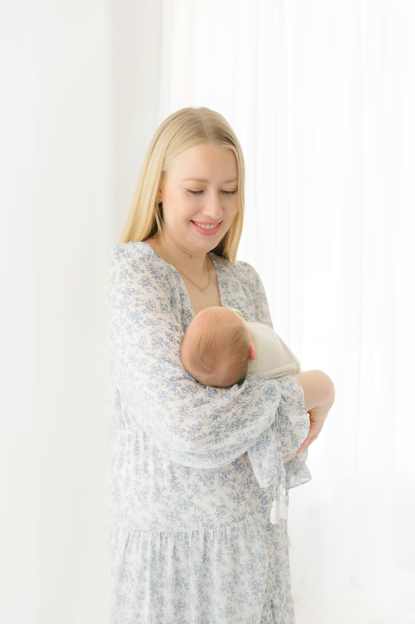 A happy new mom in a blue floral dress cradles her newborn in her arms while standing in a studio