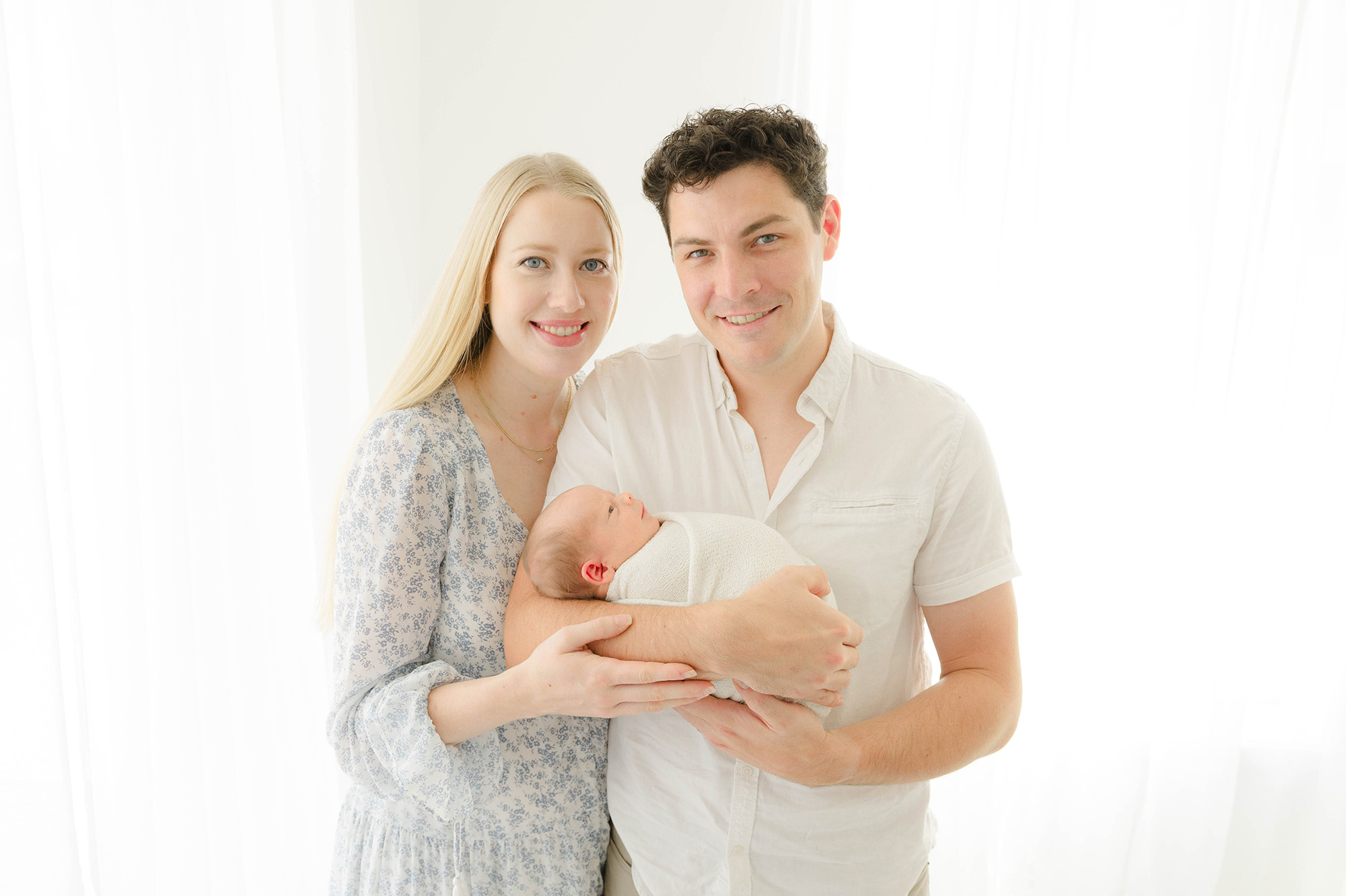 Happy new parents stand in a studio window smiling with their newborn laying in dad's arms with eyes open after leaving okc birth center