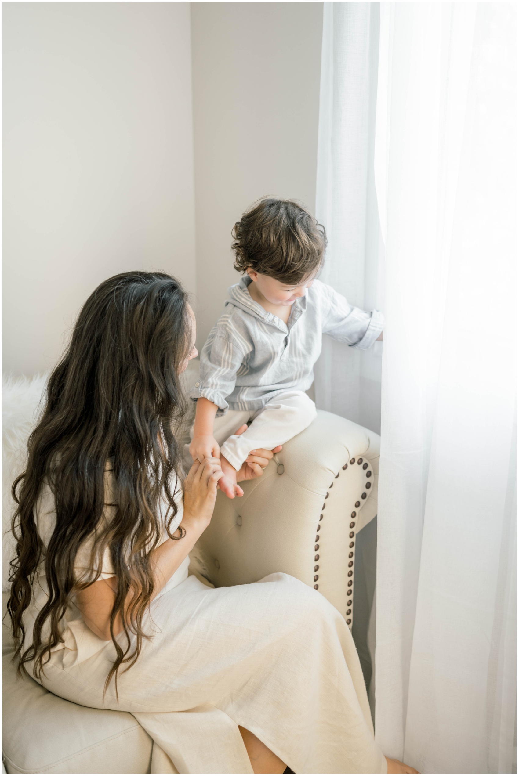 A young boy climbs on a couch arm into a window while playing with mom