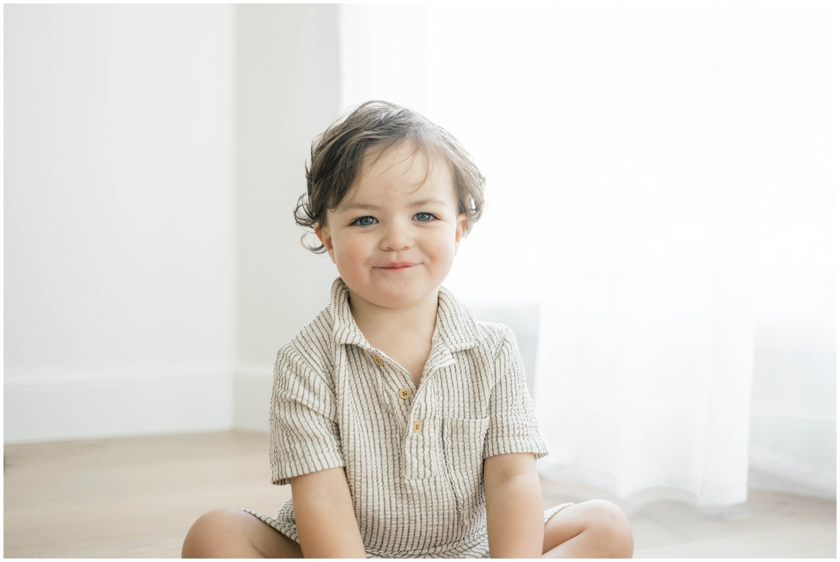 A happy toddler boy in a stripe shirt sits on the floor of a studio under a window after visiting okc clothing stores