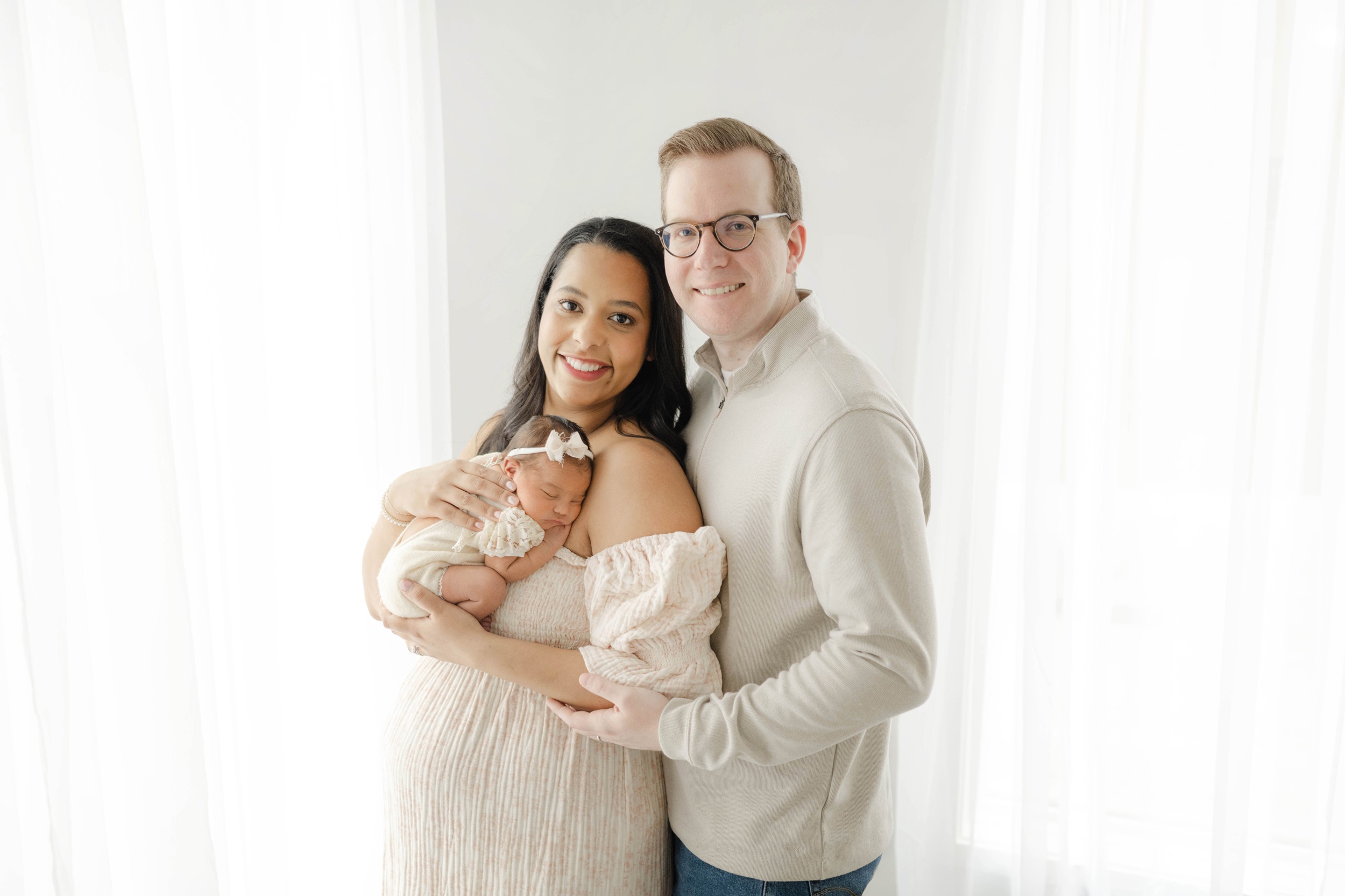 Happy new parents stand in a studio with their newborn sleeping against mom's chest after leaving ou maternal fetal medicine