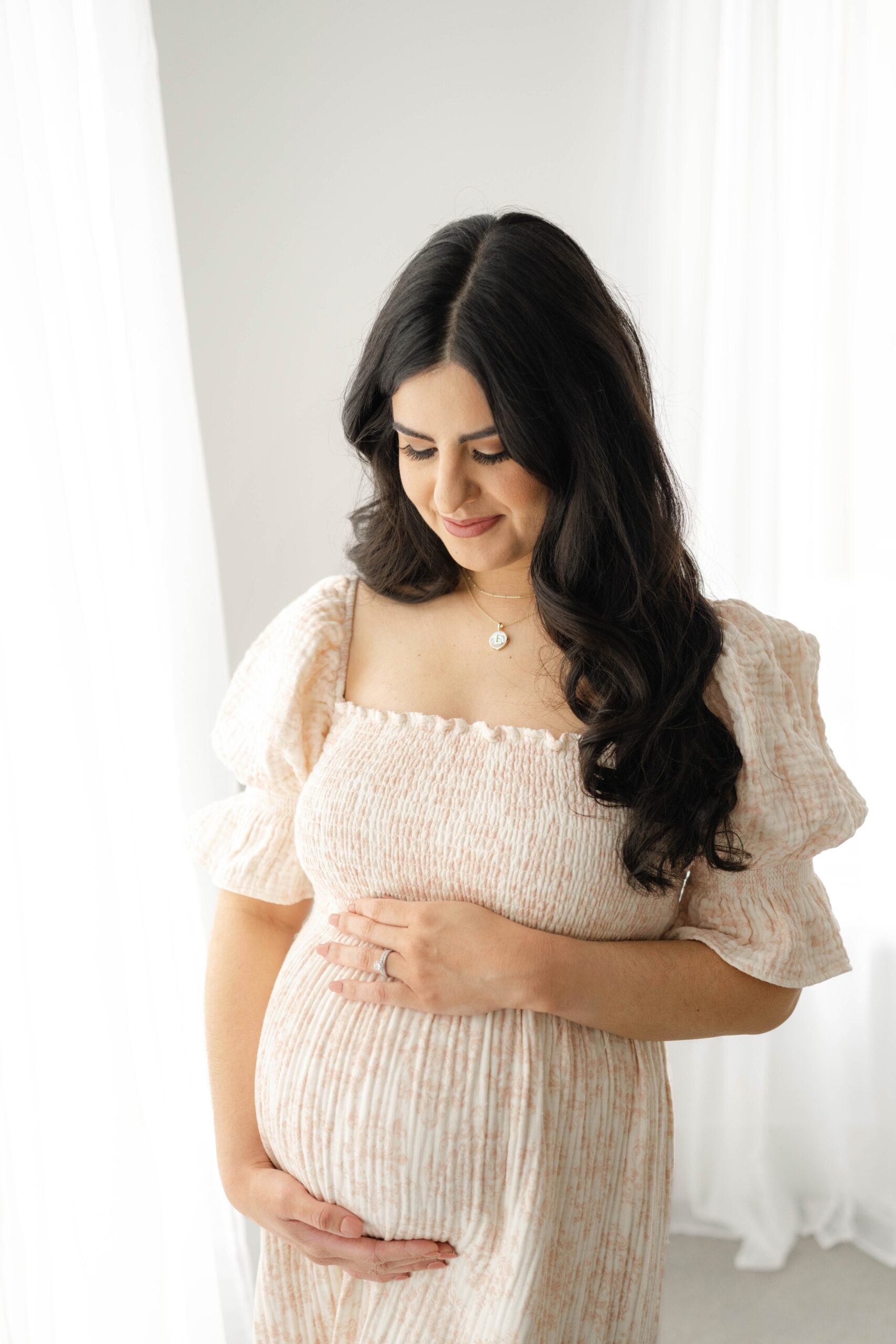 A smiling mom to be gazes down to her hands on her bump in a studio