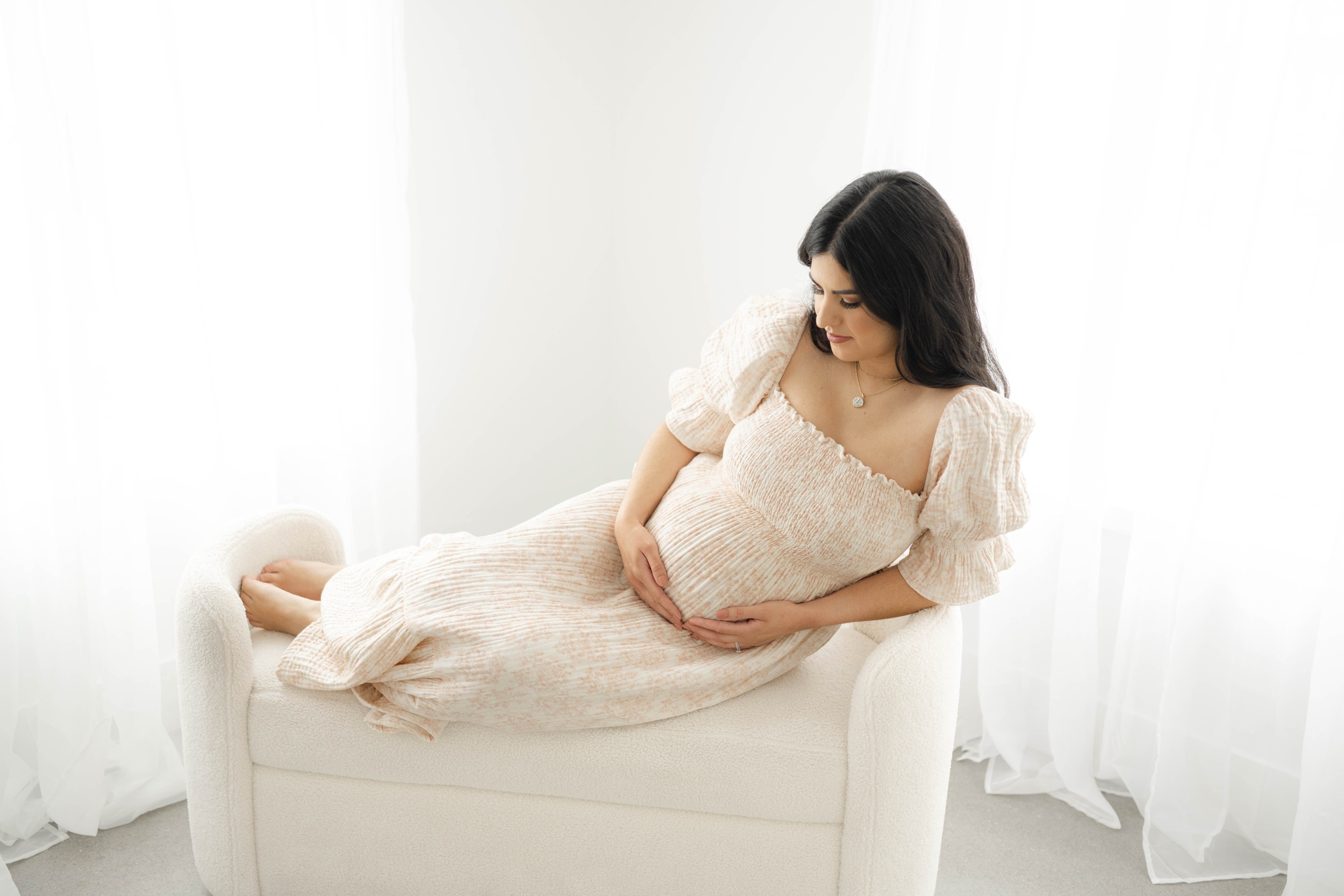 A mom to be in a pink maternity gown sits across a bench in a studio looking at her bump after visiting perinatal center okc