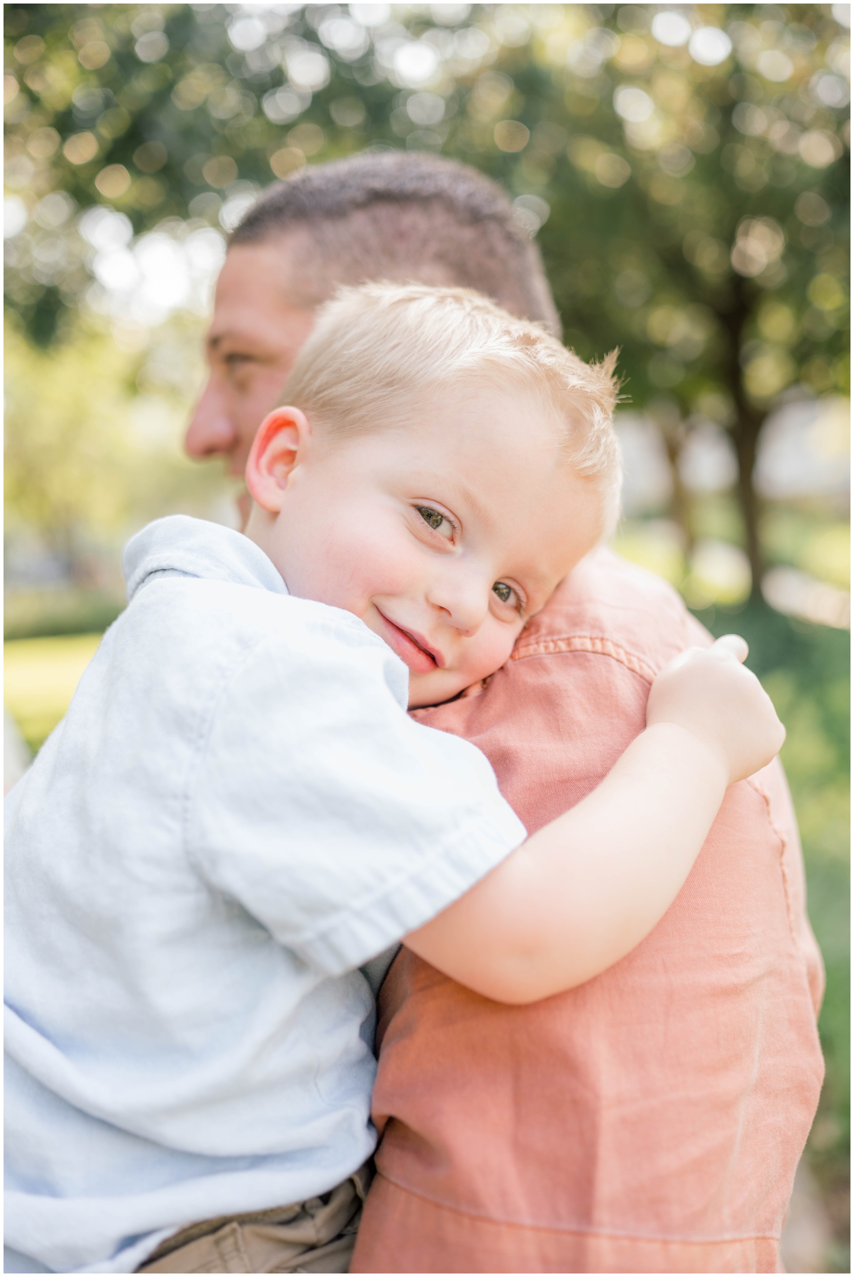 A toddler in a blue shirt leans on his dad's shoulder in a park