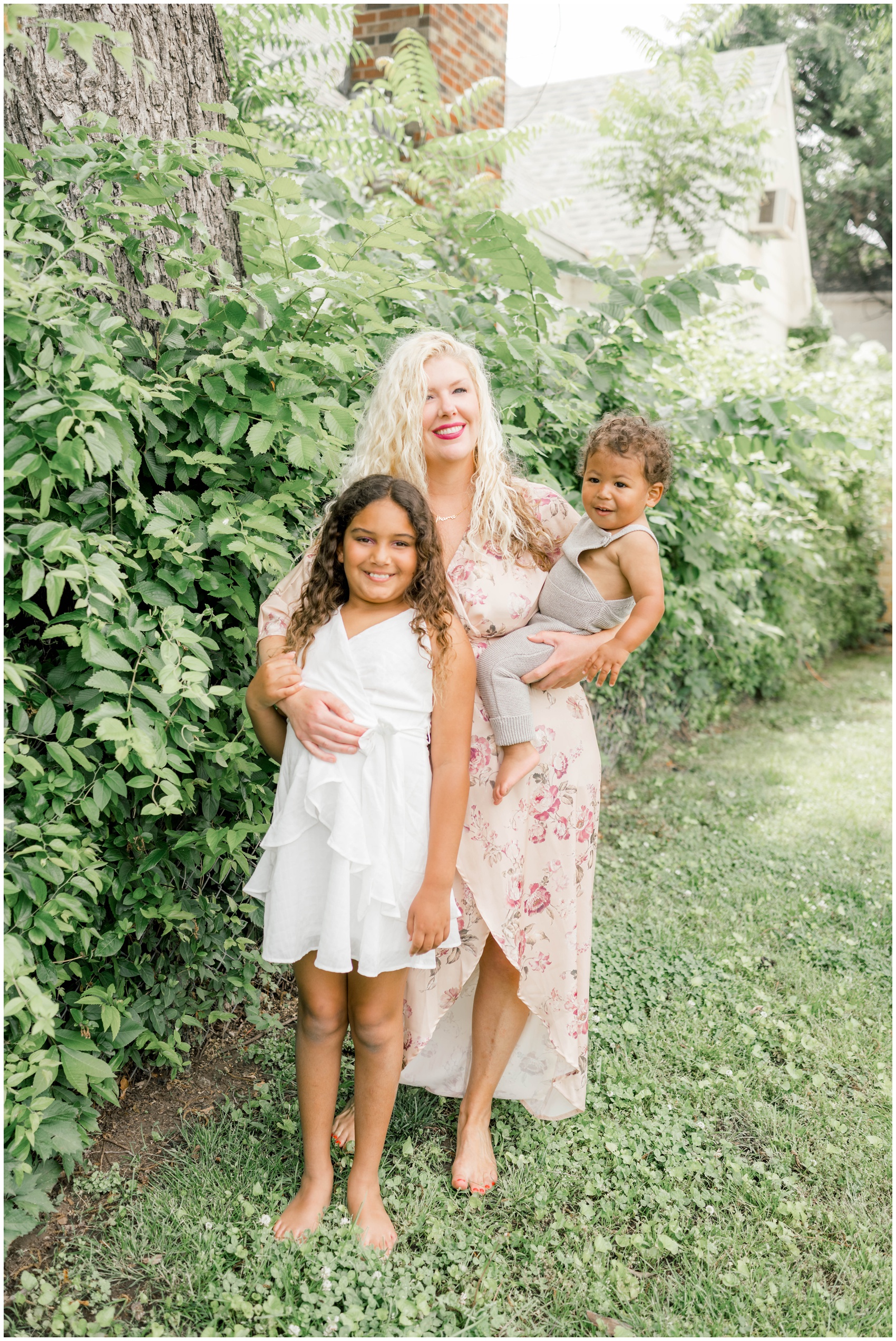 A happy mom stands in a garden barefoot with her baby on her hip and hugging her young daughter