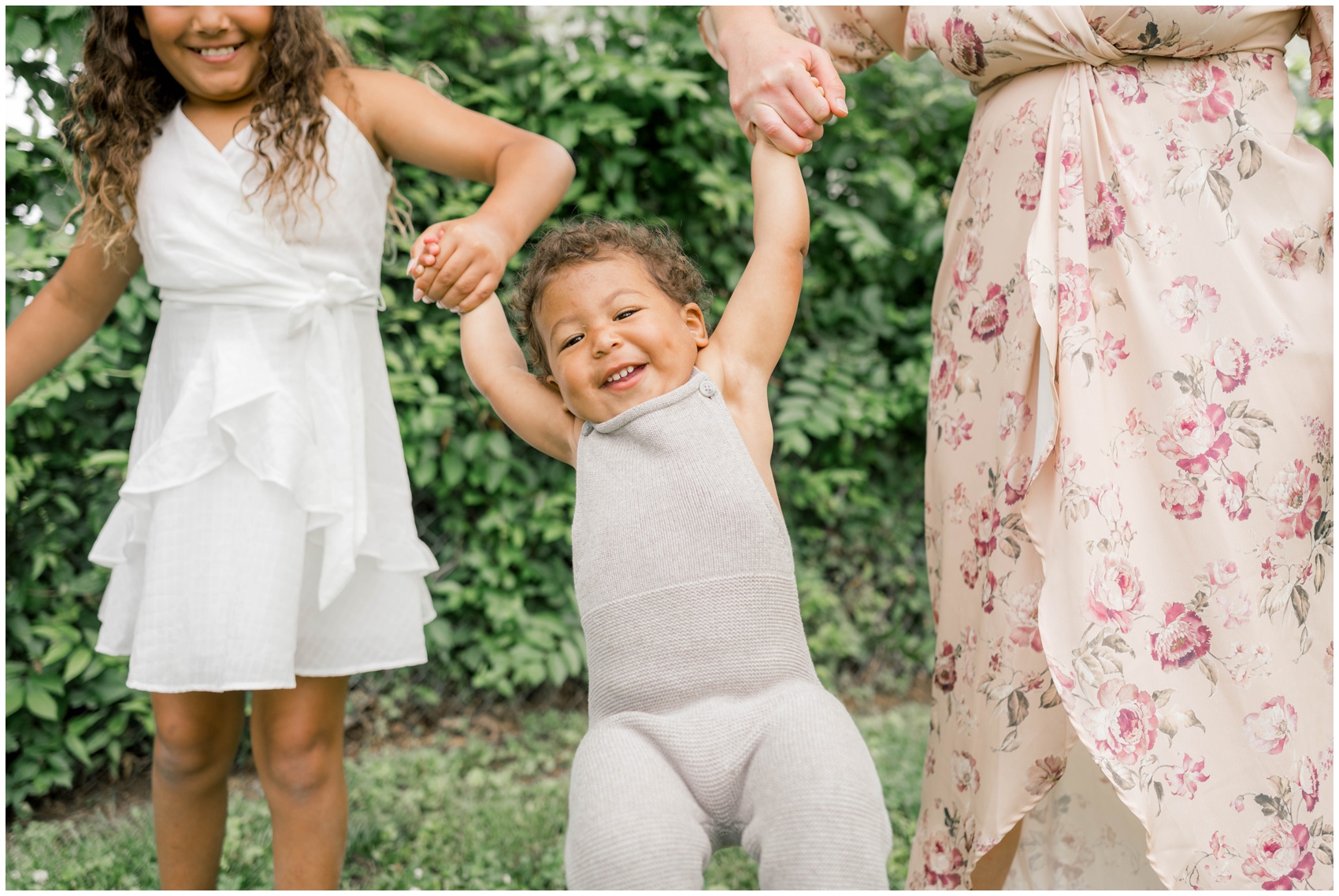 A toddler boy in a grey onesie swings in mom and big sister's hands before visiting private schools in okc