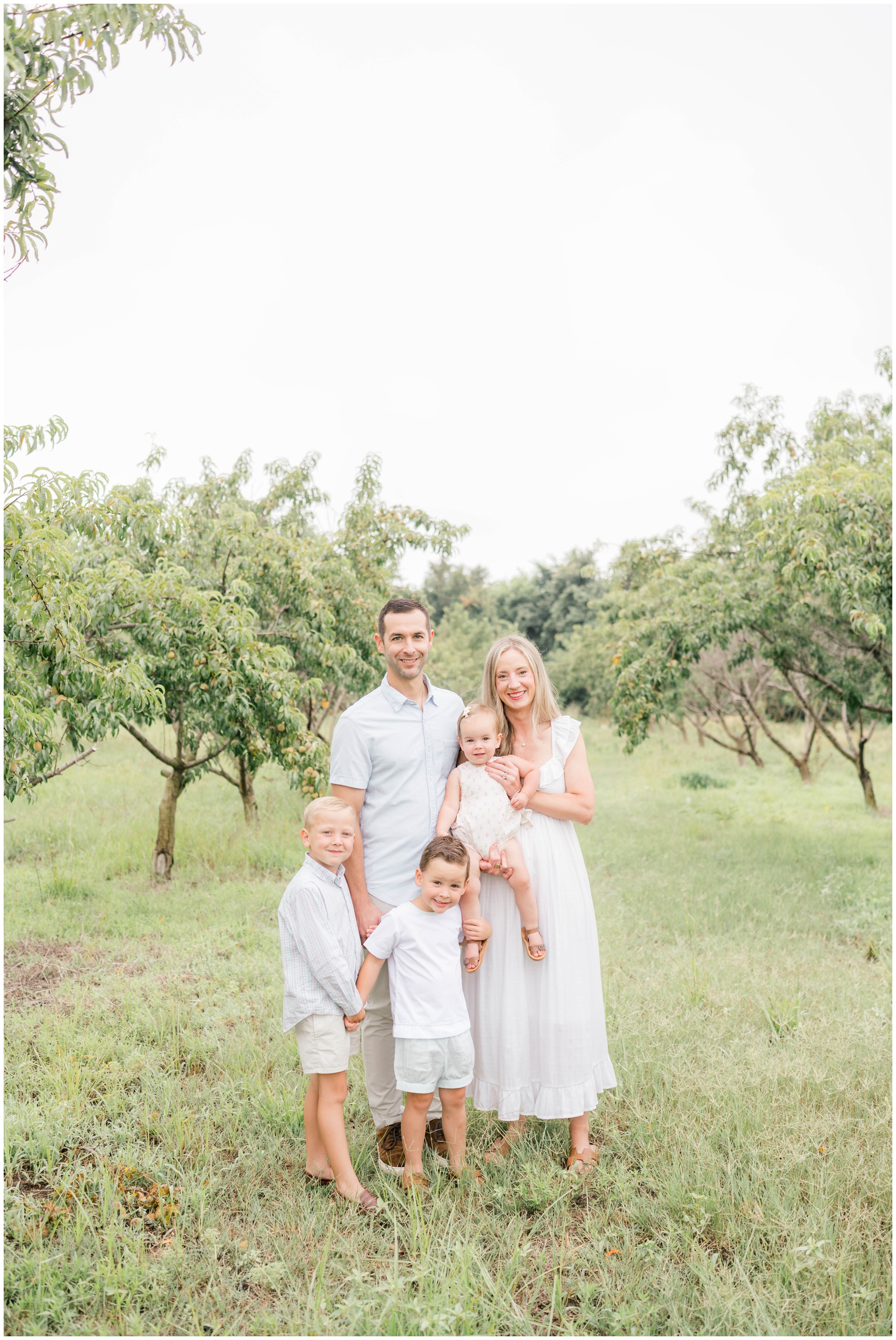 A mom and dad stand in a peach orchard with their three young children all in white before visiting pumpkin patches in okc