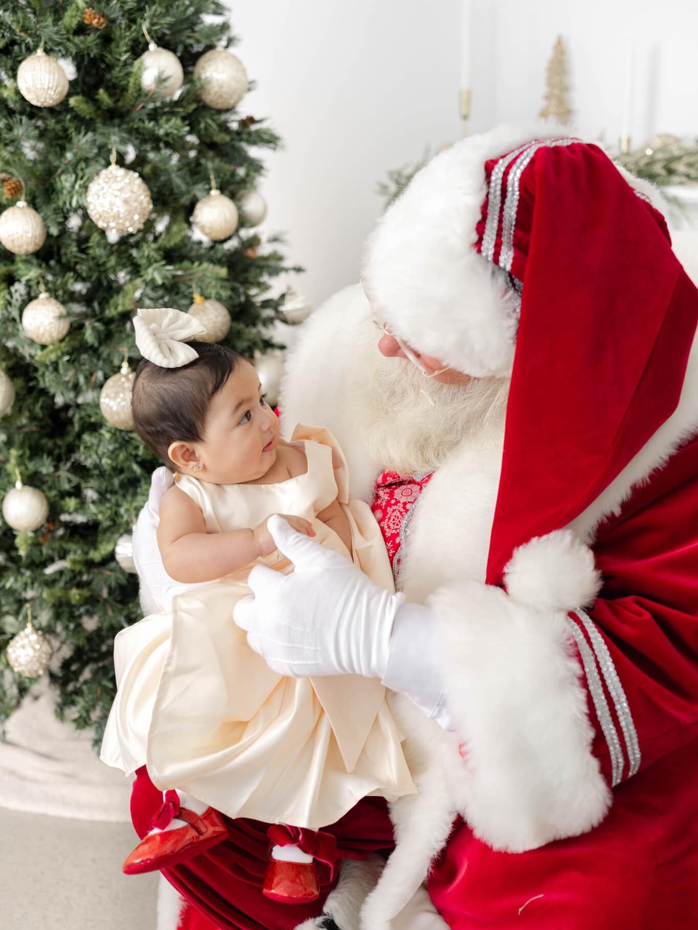 A baby girl in a white dress looks up to santa while sitting in his lap