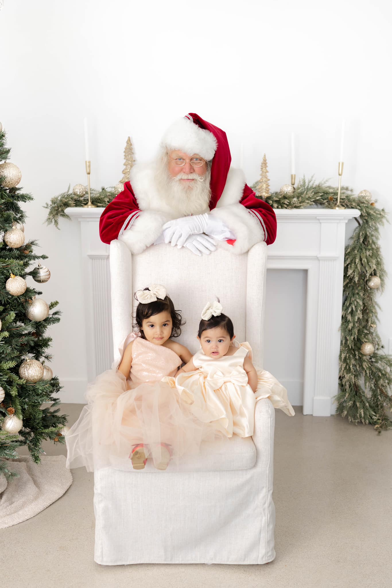 A santa in okc stands behind a chair with toddler sisters sitting in it by a tree and fireplace