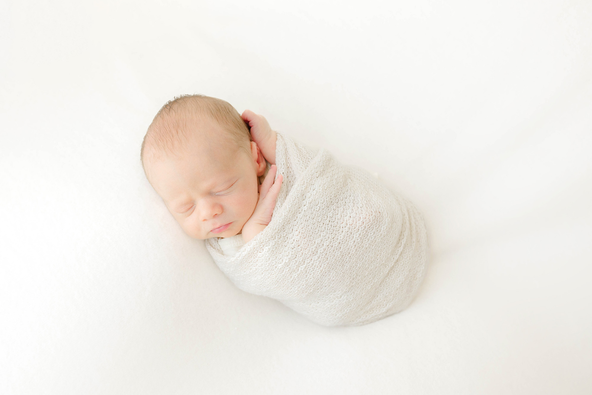 A newborn sleeps in a tight white swaddle on a white bed thanks to a sleep coach okc