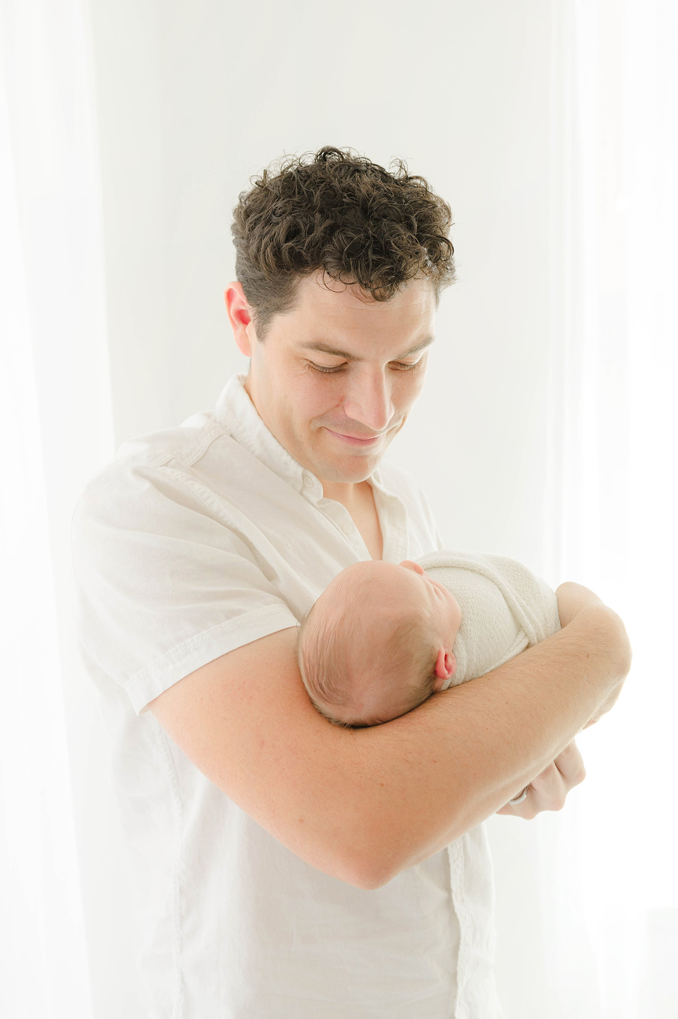 A new dad smiles down at his sleeping newborn in his arms in a studio window thanks to a sleep coach okc