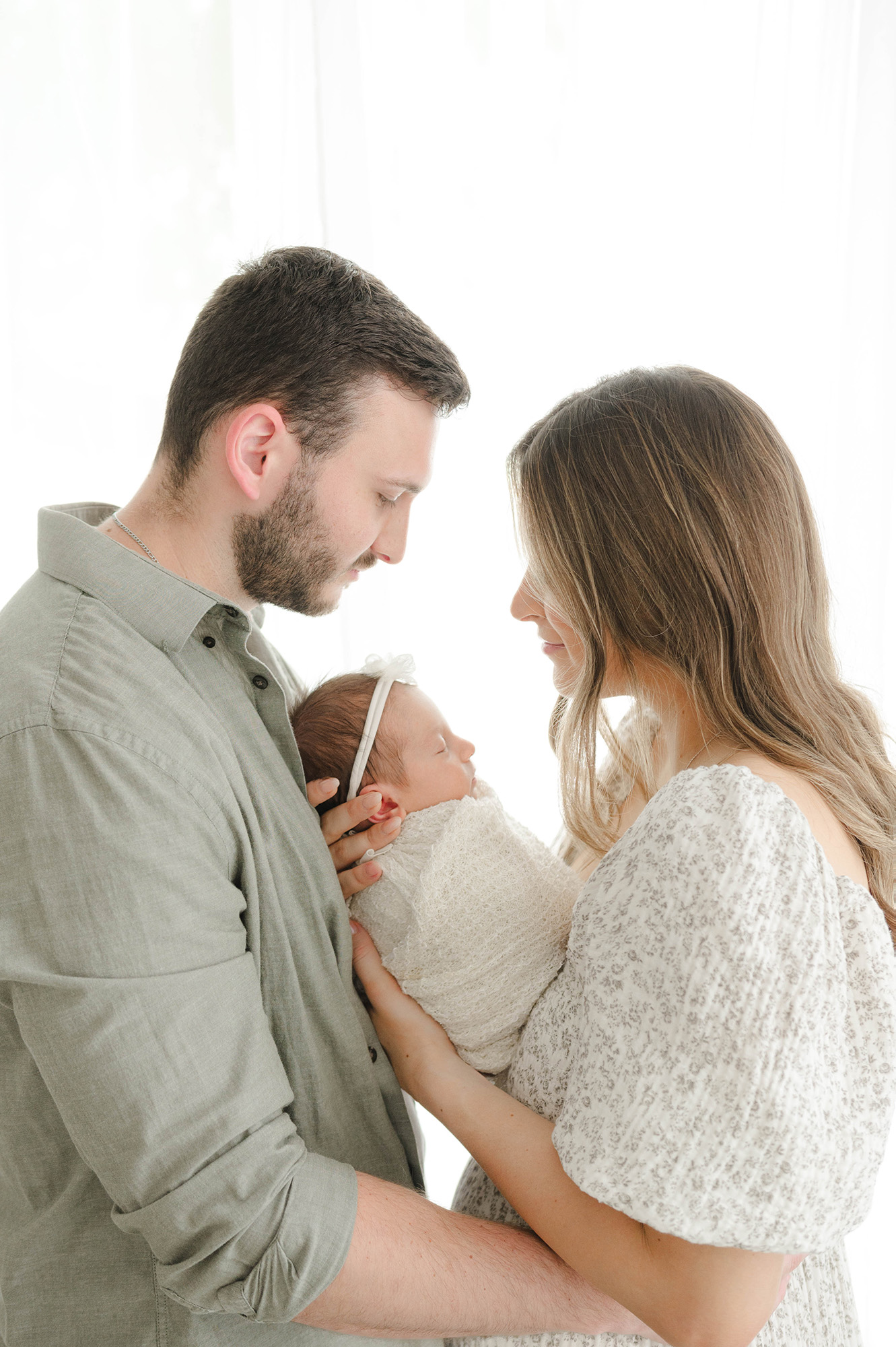 Happy new parents stand in a window with sleeping newborn daughter between them thanks to spirited birth midwifery