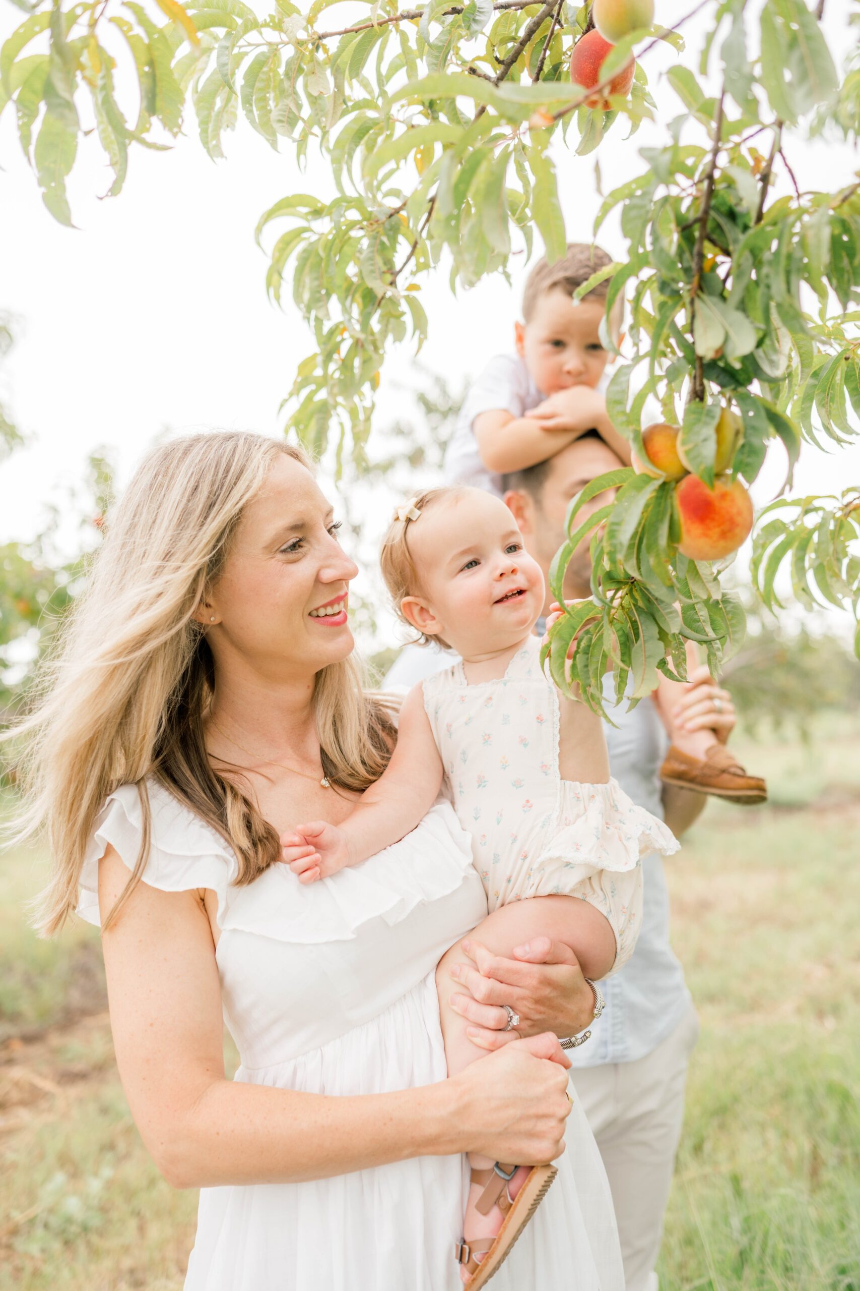 A happy mom and dad explore a peach orchard with their toddlers on hip and shoulders before some spring break camps okc