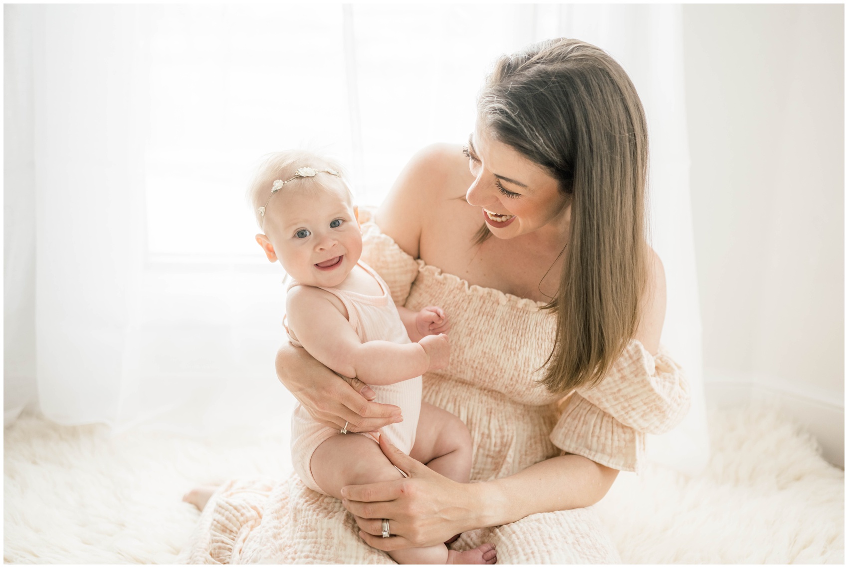 A mom and baby daughter laugh while playing in mom's lap on a rug under a window