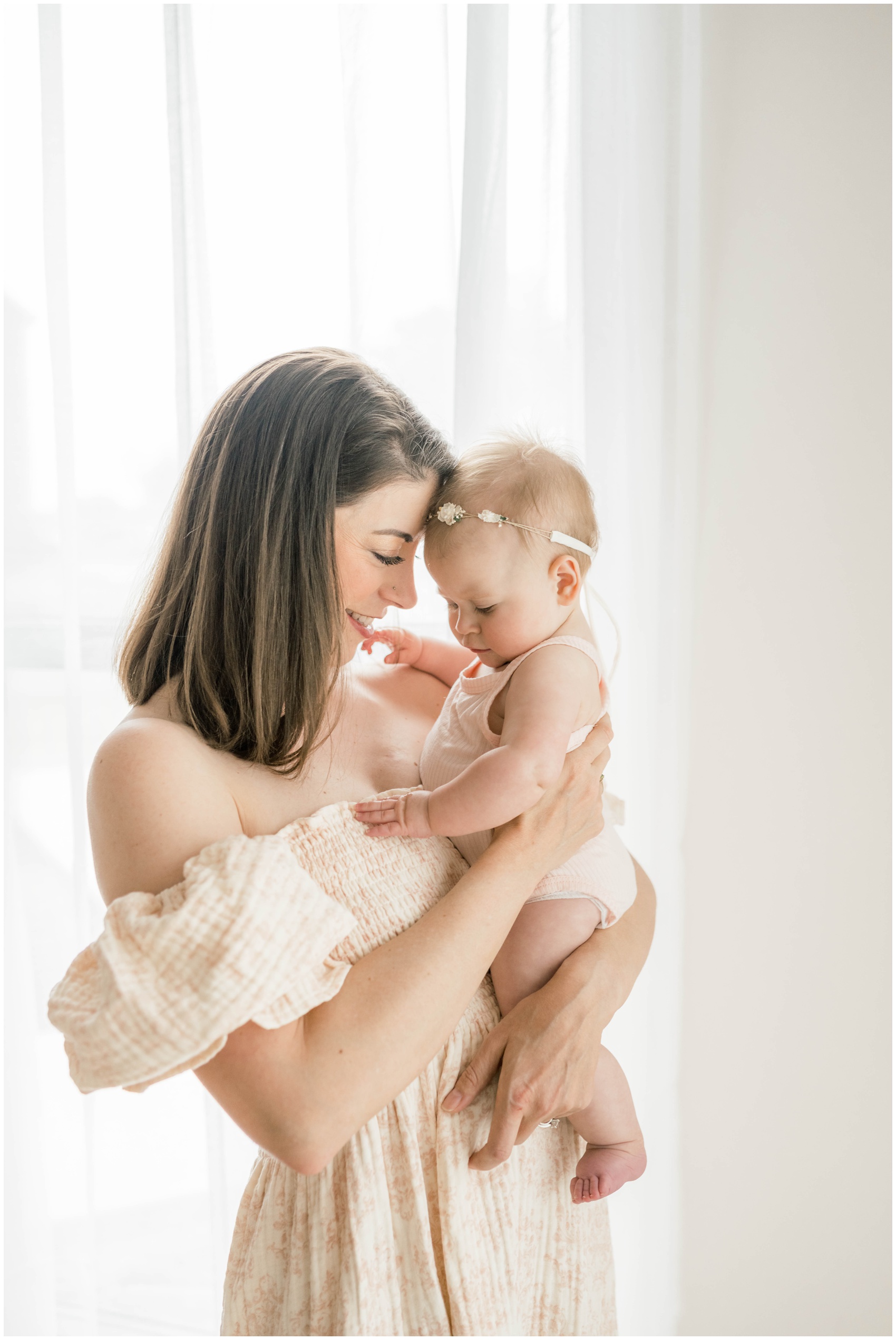 A happy mom in a cream dress snuggles her baby daughter in her arms while standing in a window before some swim lessons in okc