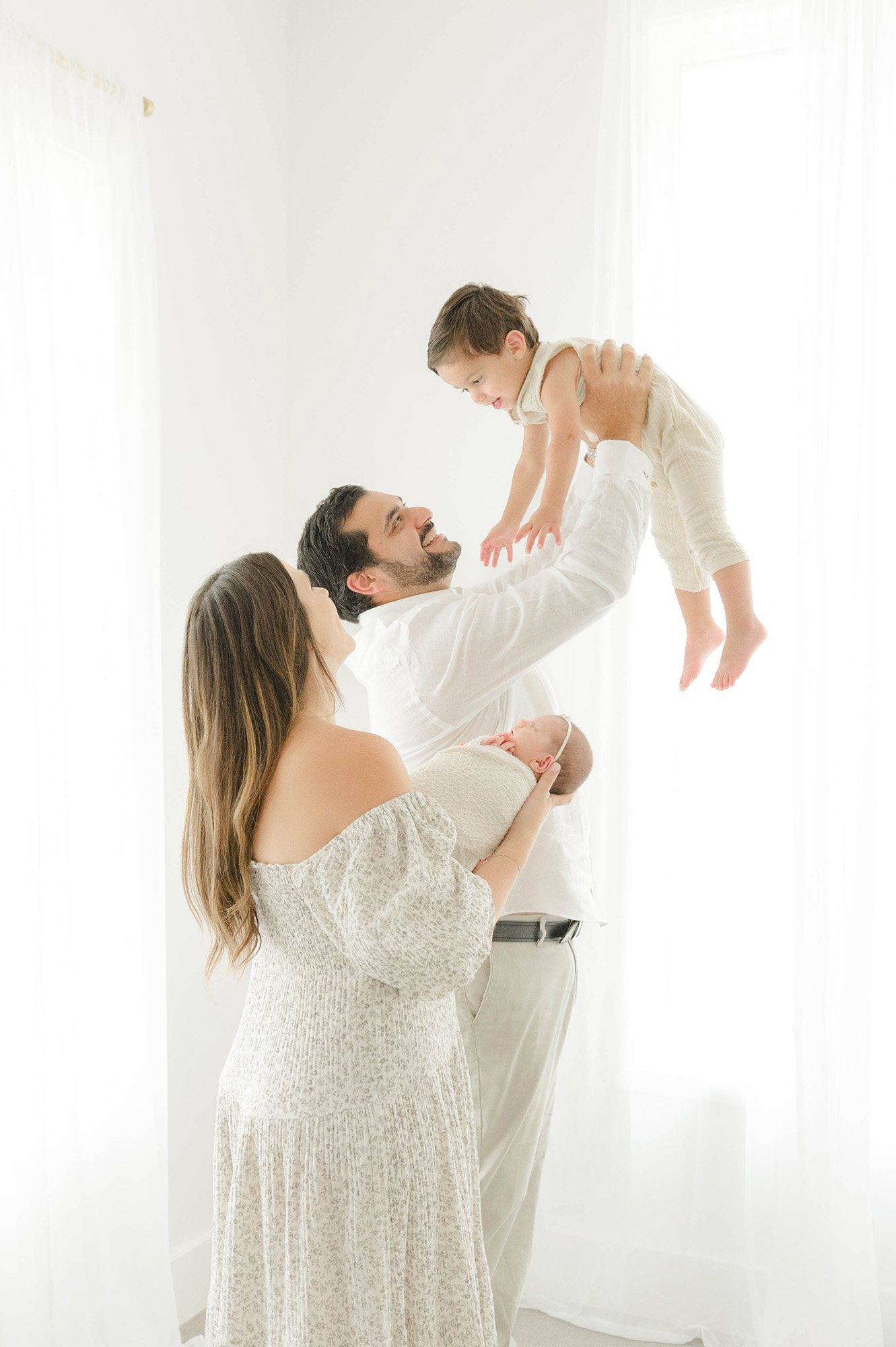 A happy dad lifts his giggling toddler above his head while mom holds a newborn