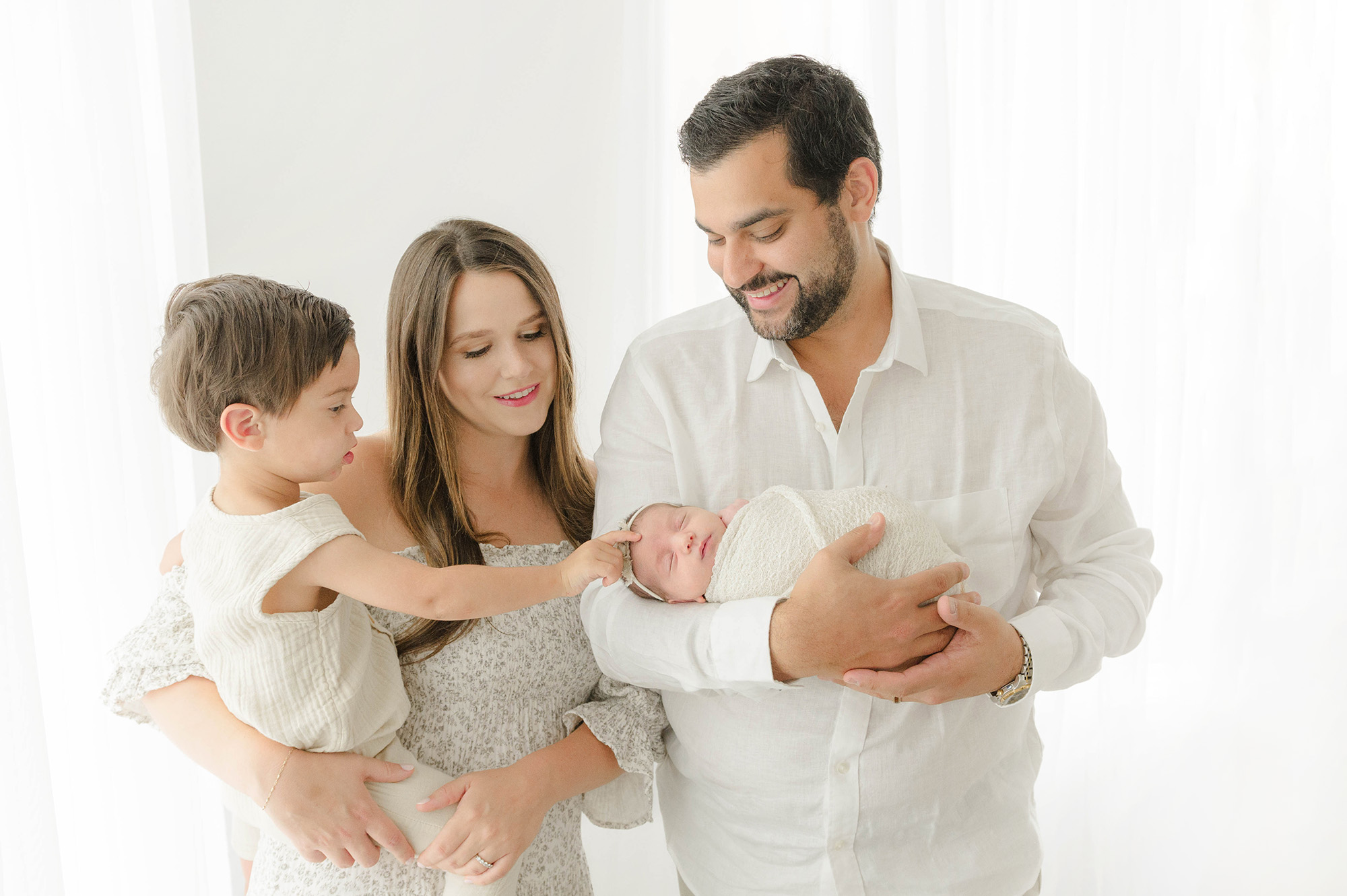 A toddler boy touches his sleeping baby sister's head in mom and dad's arms in a studio during things to do with kids in oklahoma city