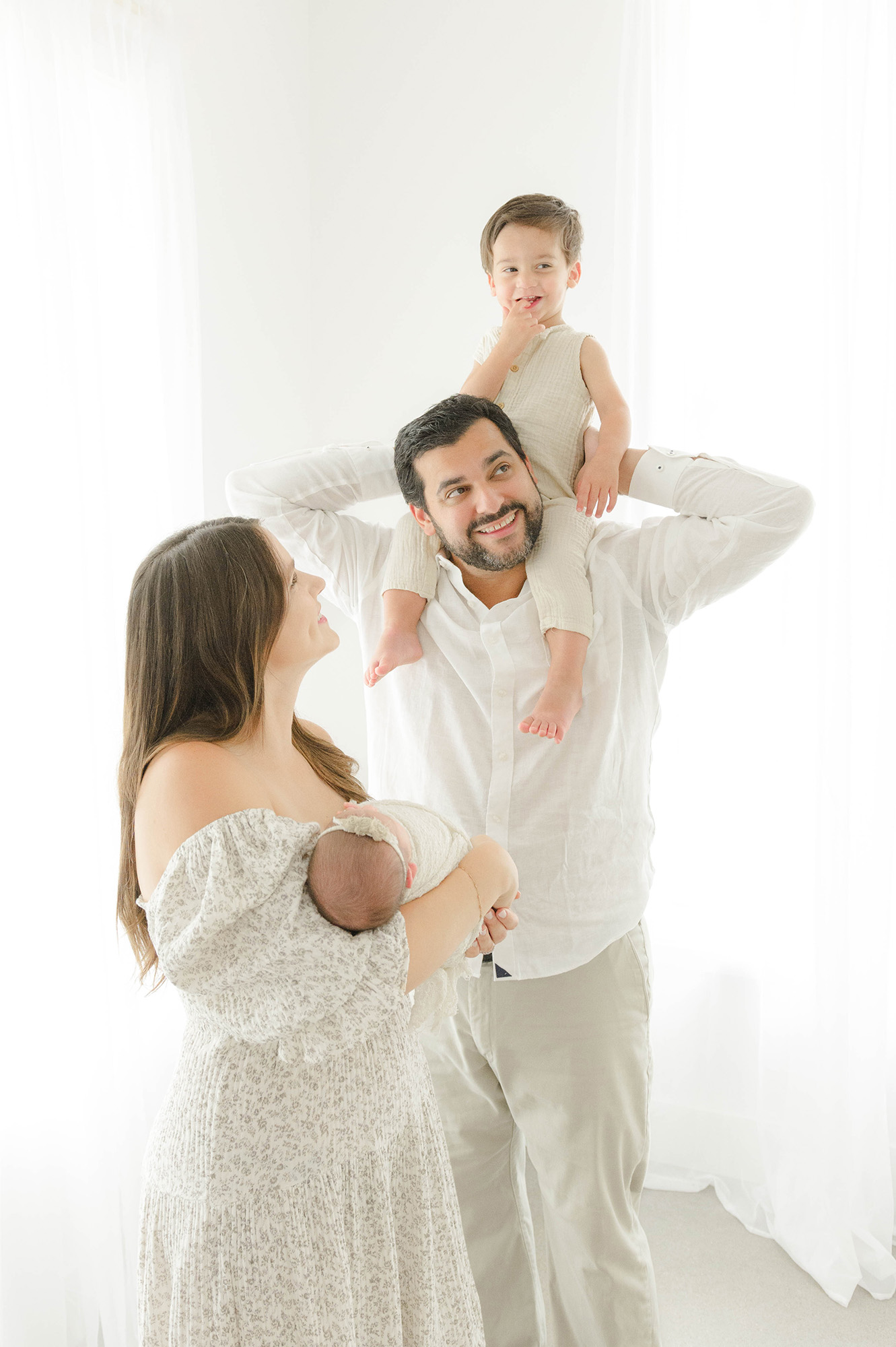 A happy toddler boy sits on dad's shoulders in a studio while mom holds a newborn and smiles during things to do with kids in oklahoma city