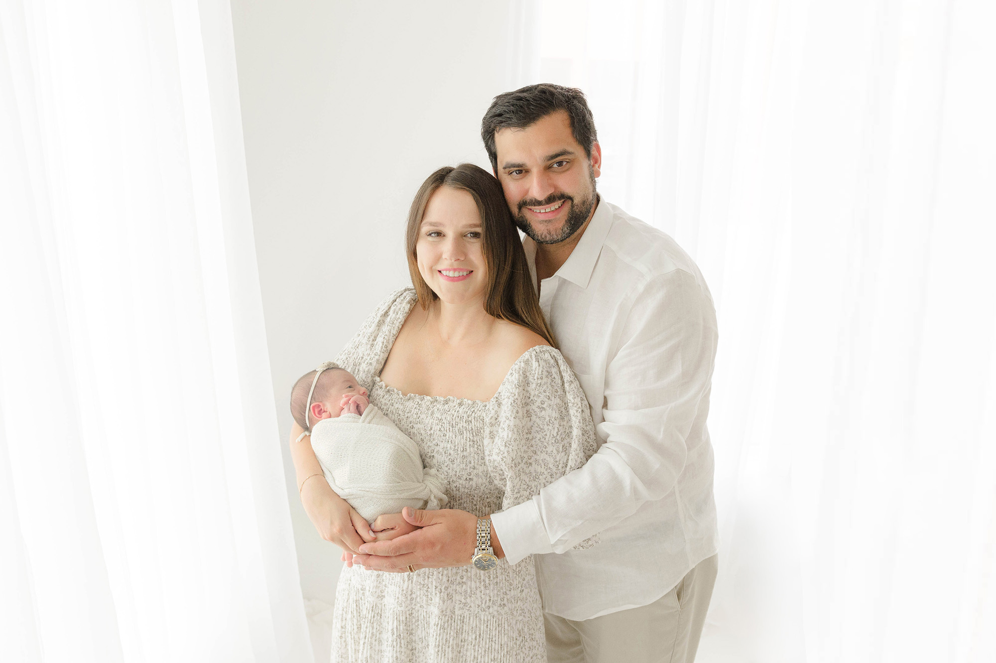 A mom and dad hug and stand in a studio holding their newborn baby daughter before visiting a tongue tie specialists in oklahoma city