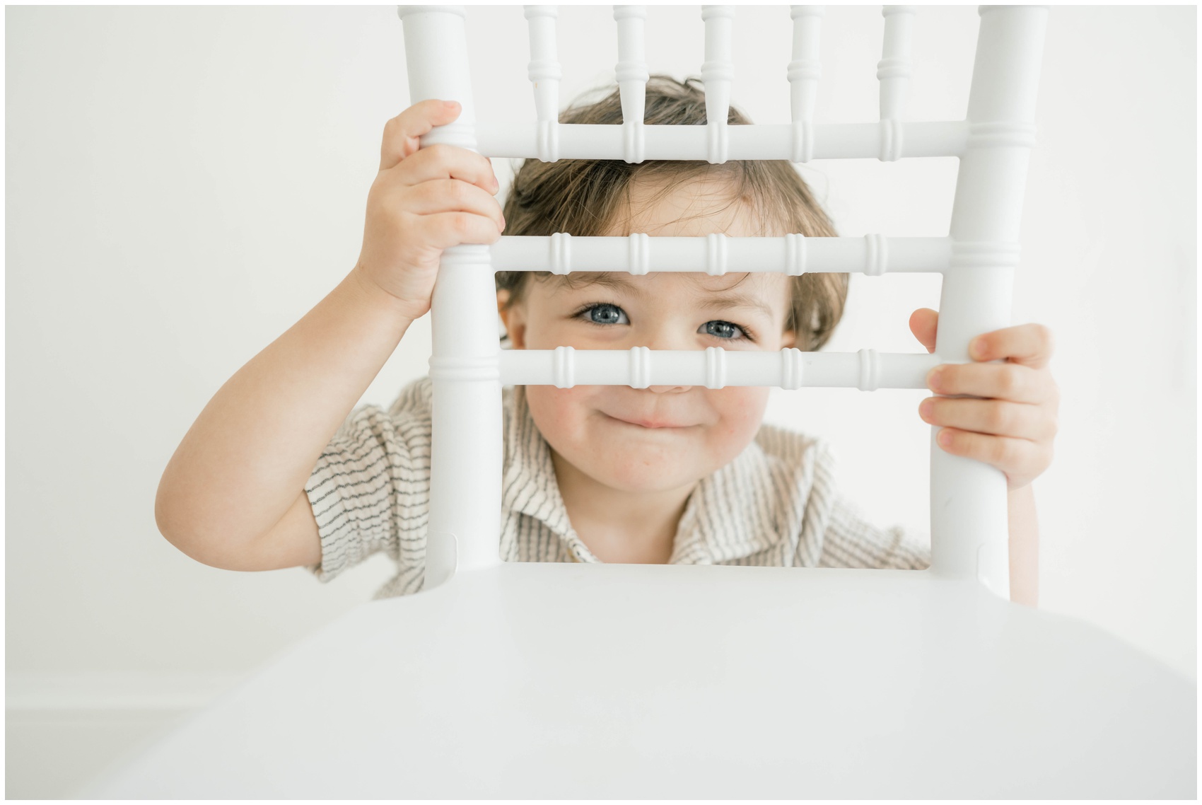 A toddler boy peeks through the back of a white wooden chair while smiling