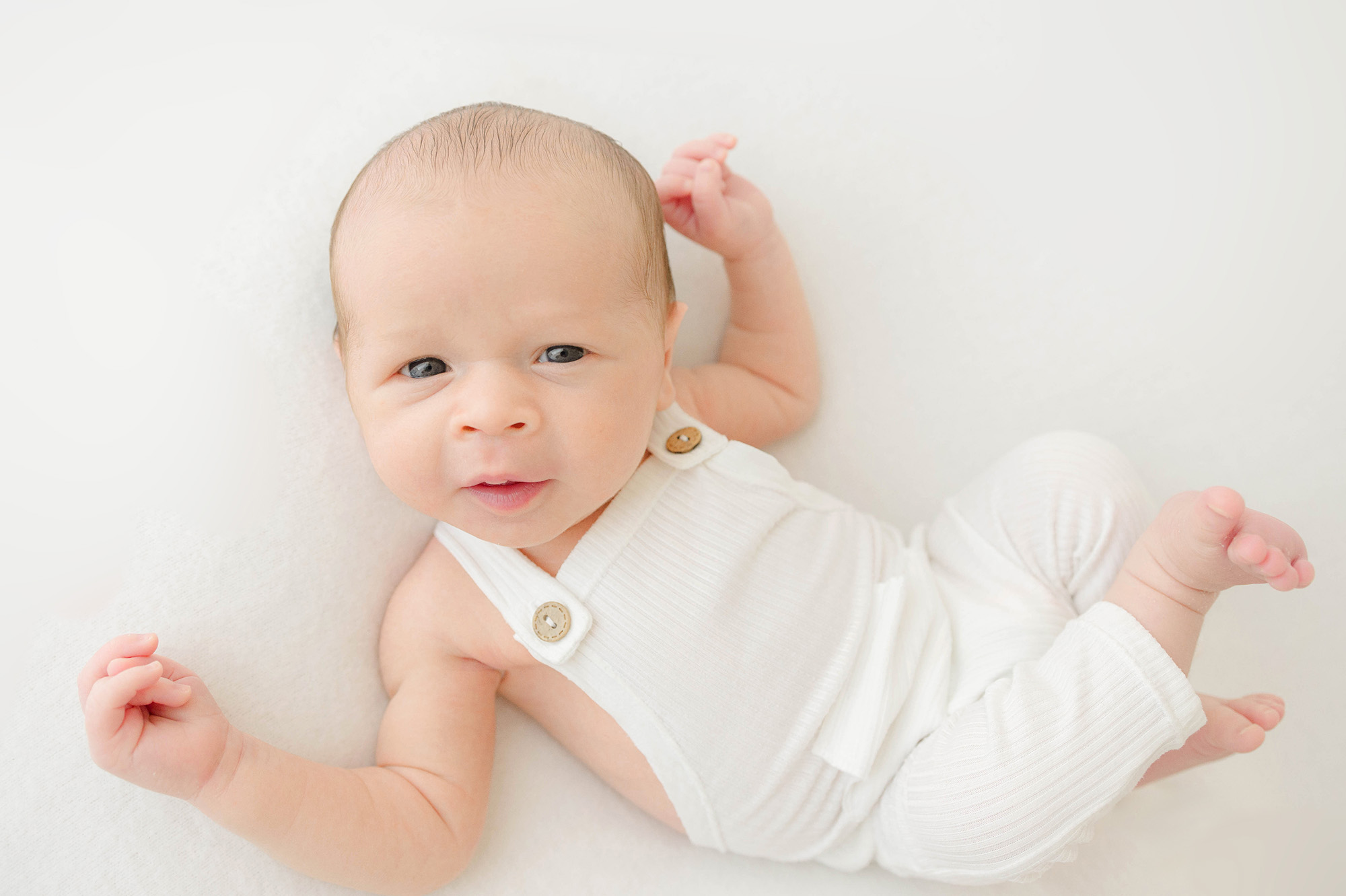 A baby smiles with eyes open while laying in white overalls